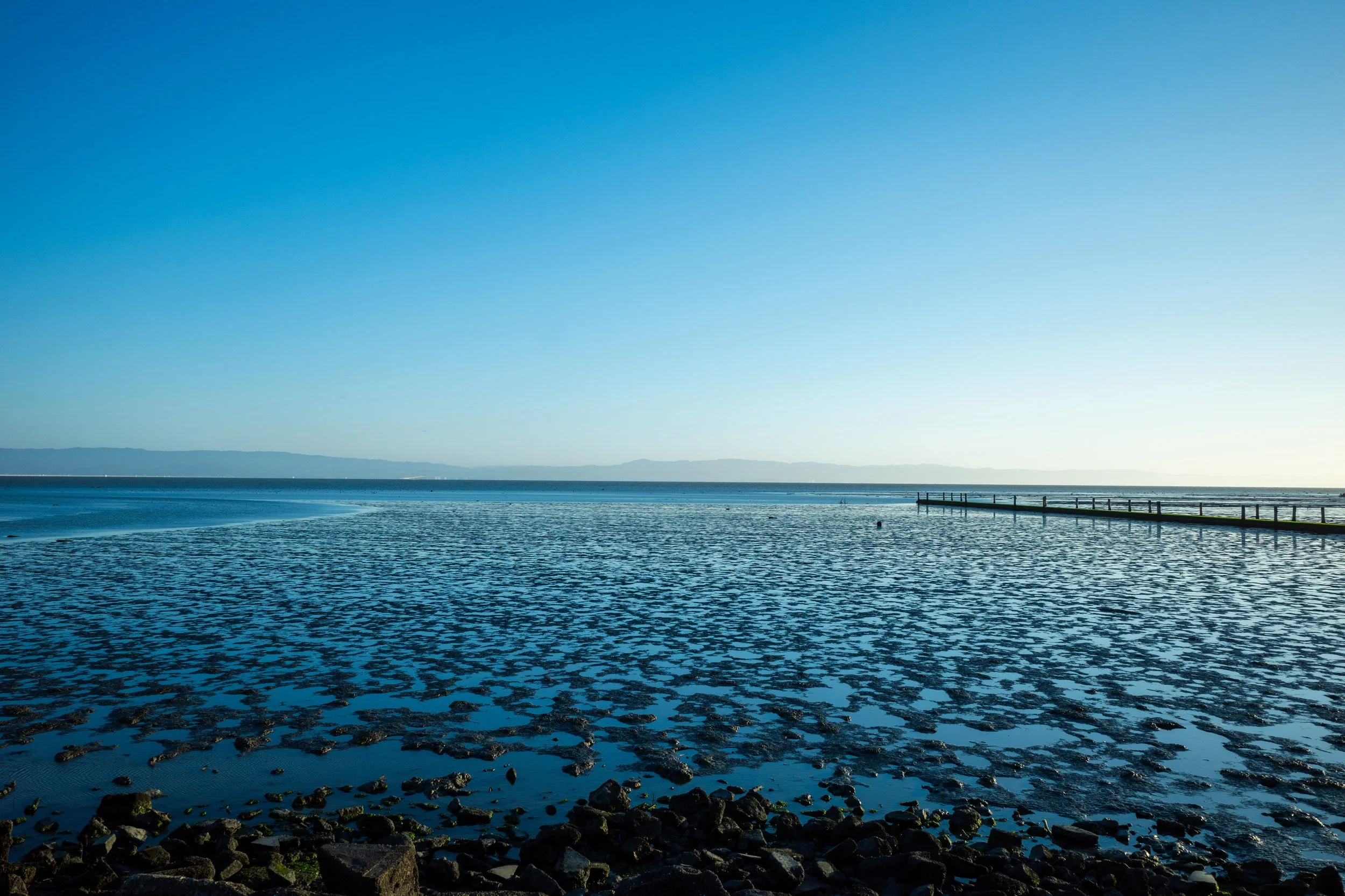 A calm body of water with rocks along the shoreline, a pier extending into the water, and mountains in the distance under a clear blue sky.