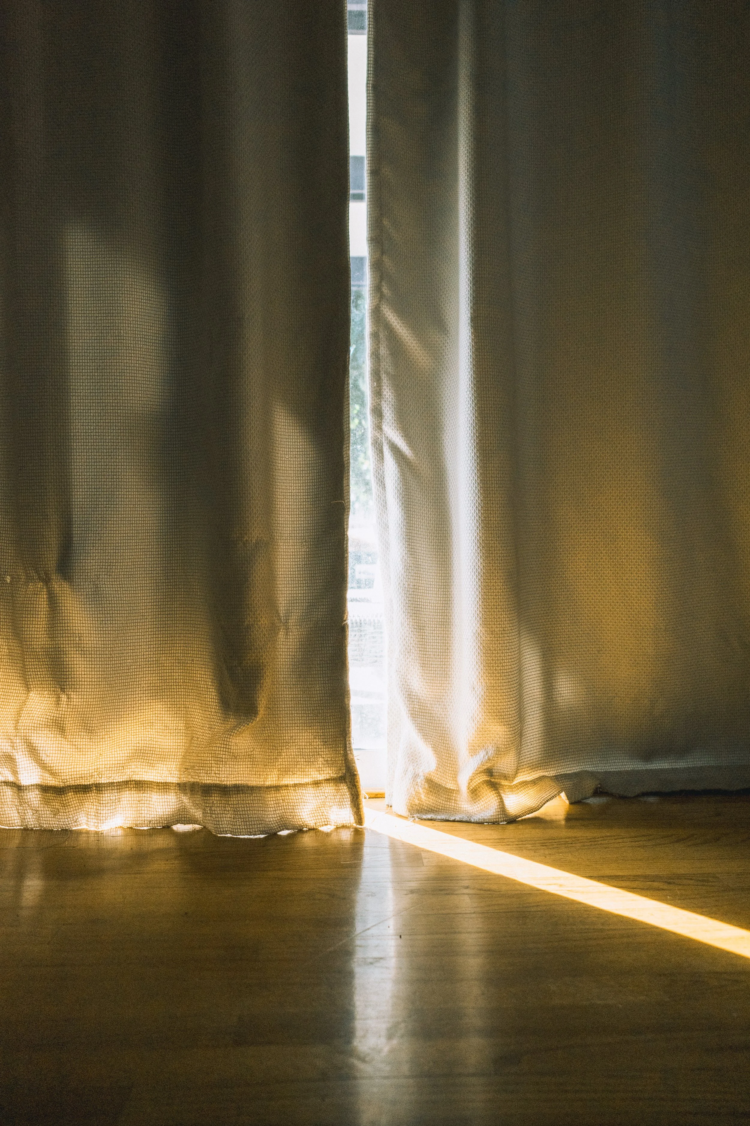 Sunlight shines through slightly parted beige curtains onto a hardwood floor.