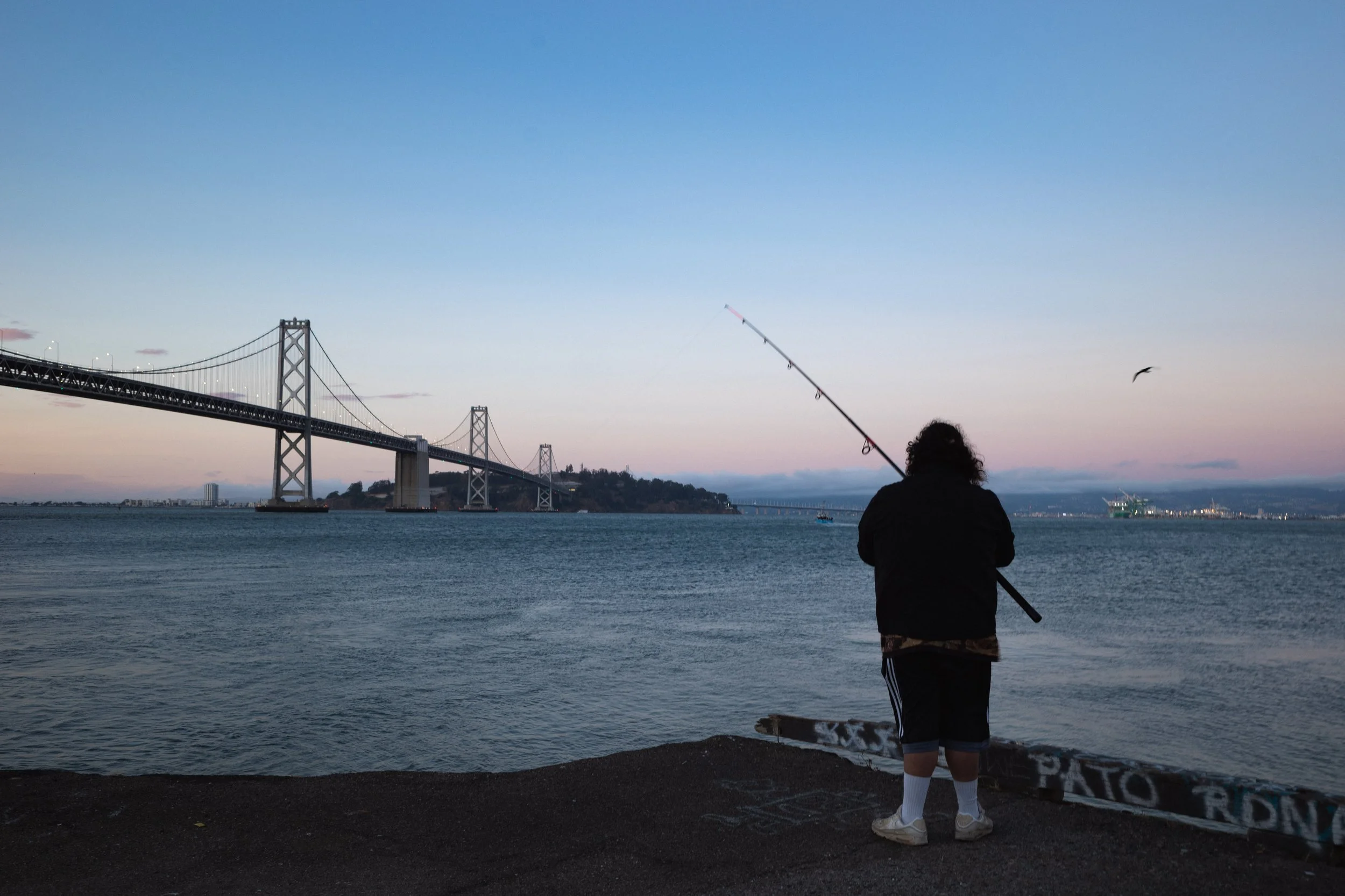 Person fishing by the water at dusk with the San Francisco-Oakland Bay Bridge in the background.