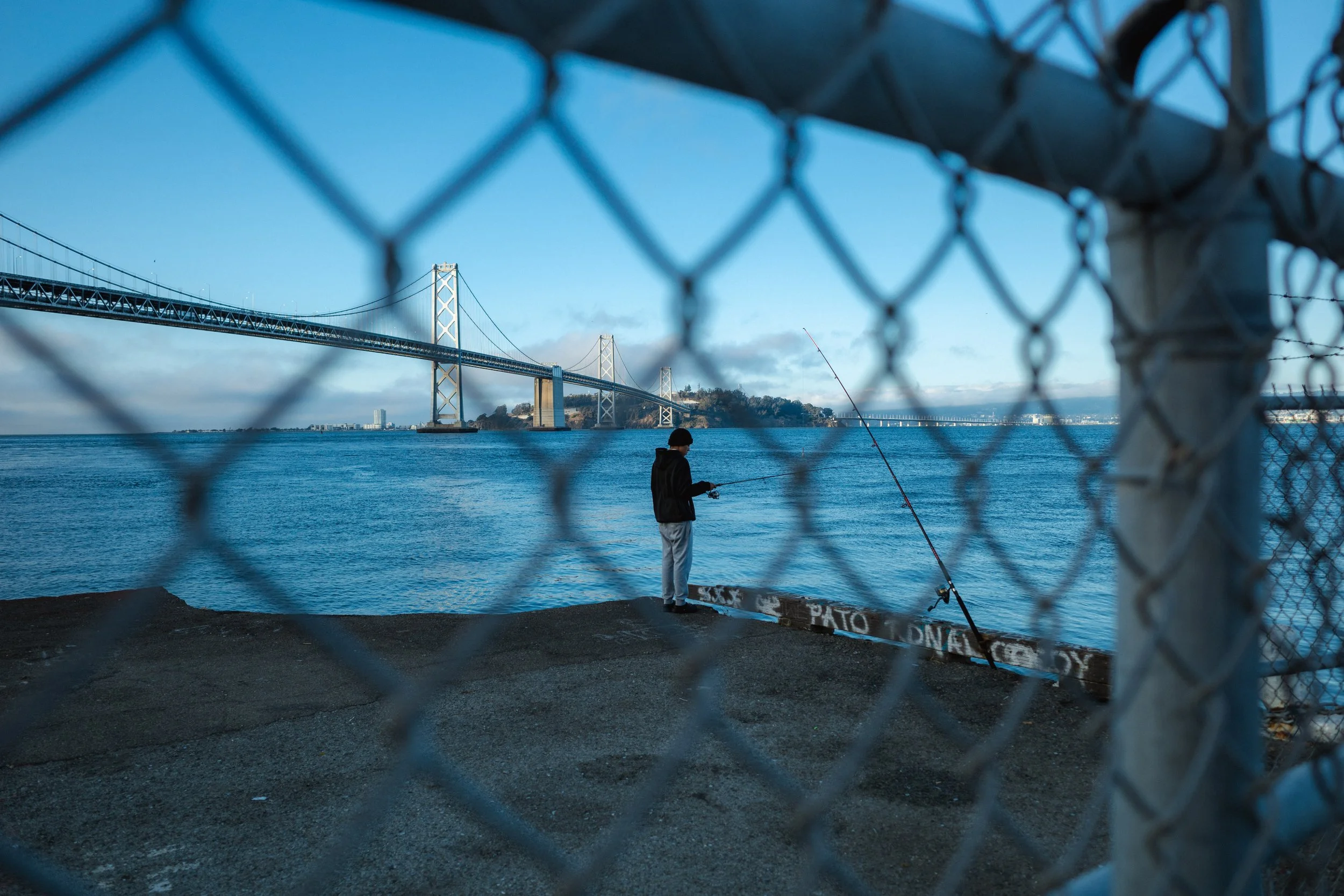 A person fishing by the water, seen through a chain-link fence, with the San Francisco Bay Bridge in the background under a partly cloudy sky.