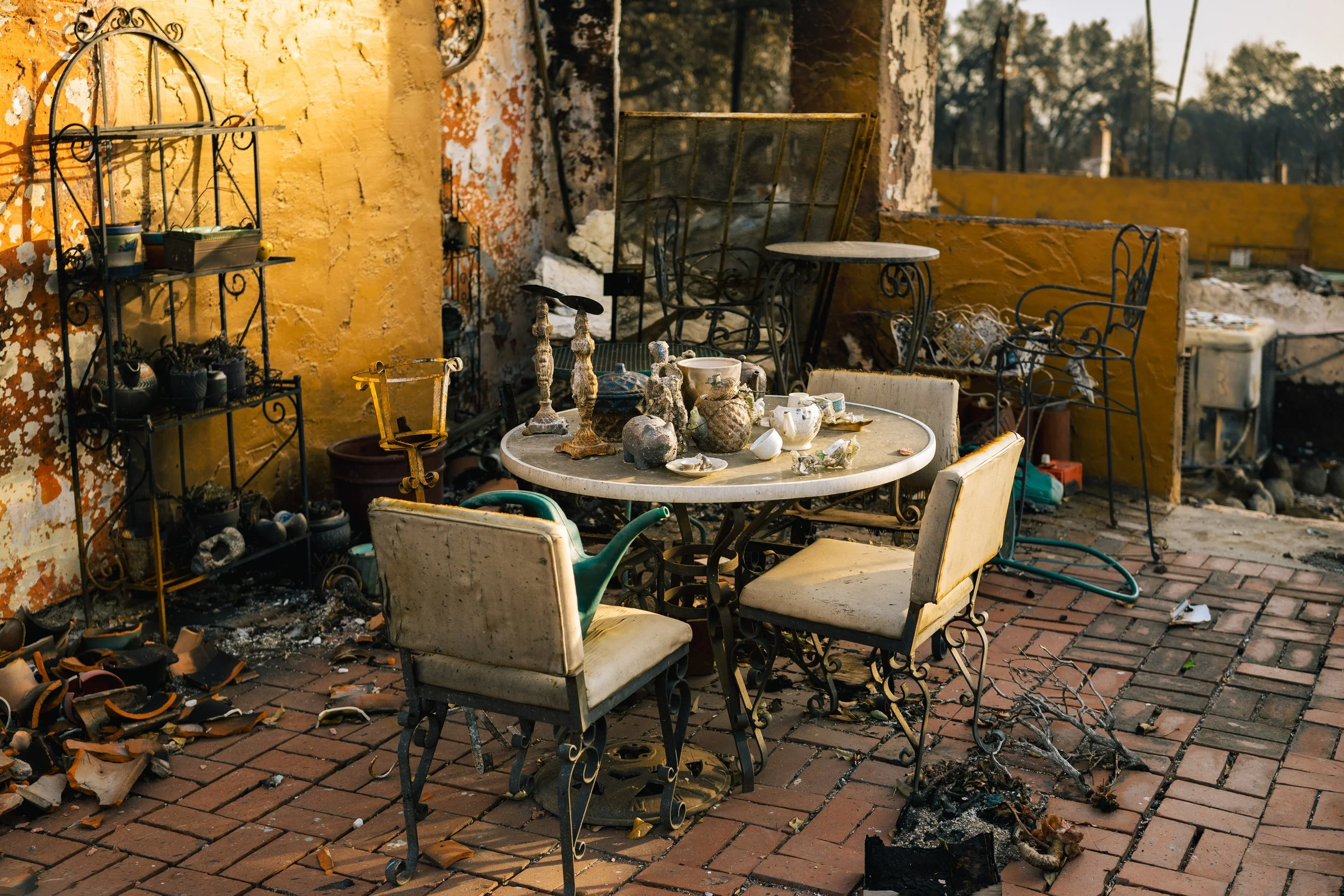 Outdoor patio area with smoking or burned furniture, charred debris, and burnt items on a brick floor. There are tables and chairs, some covered in dirt, with a yellow wall in the background and a damaged fence.