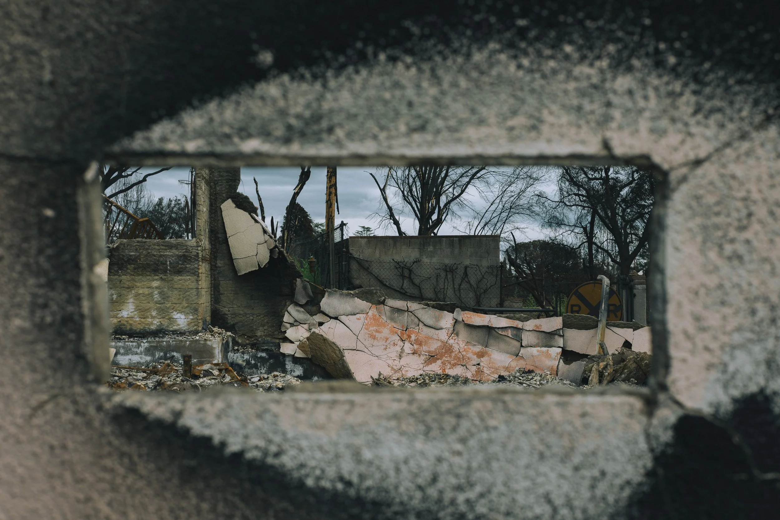 View of destruction through a small opening in a concrete wall, showing fallen debris, damaged structures, and trees with overcast sky in the background.