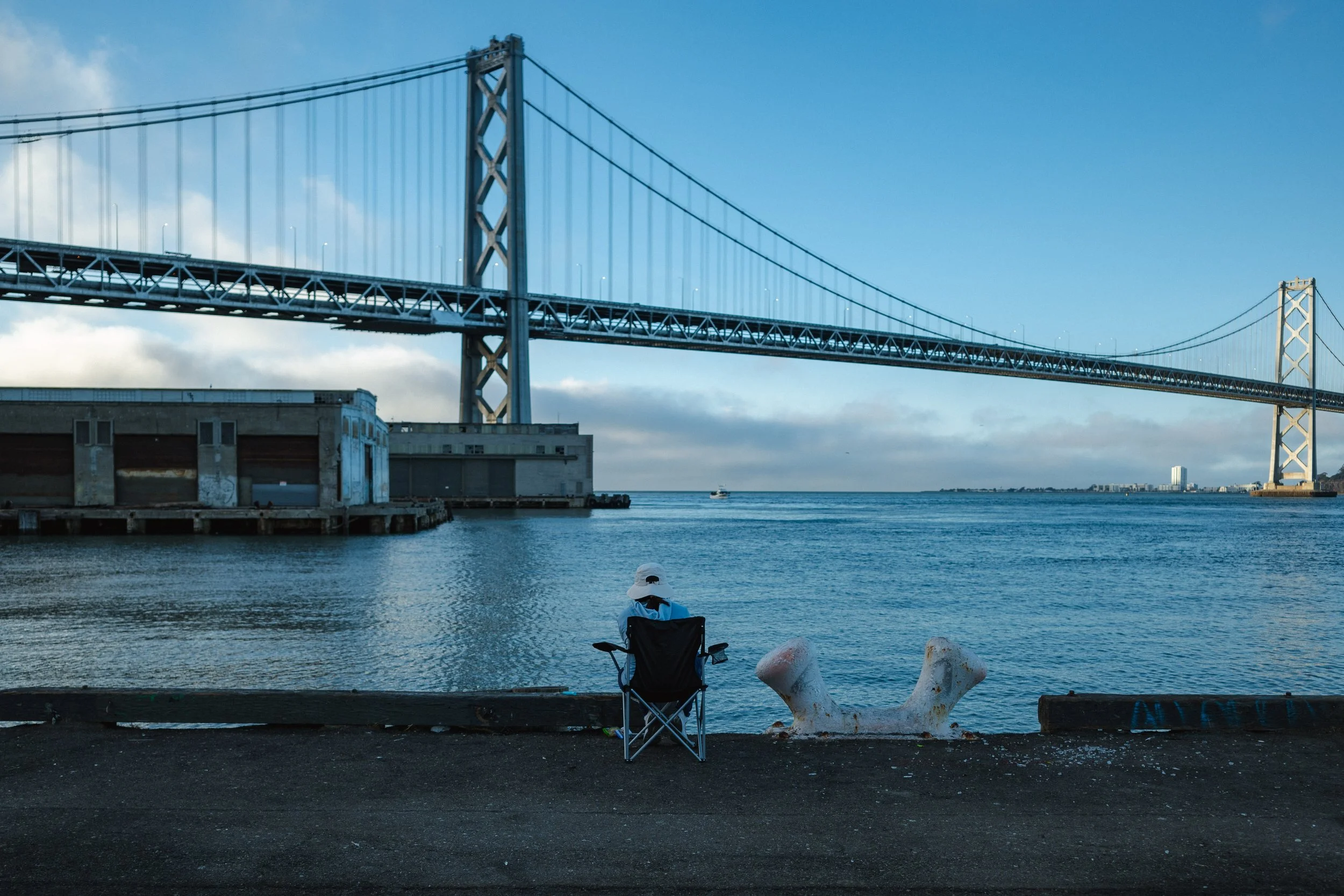Person sitting on a folding chair by the water with a large bridge in the background and a boat on the water.