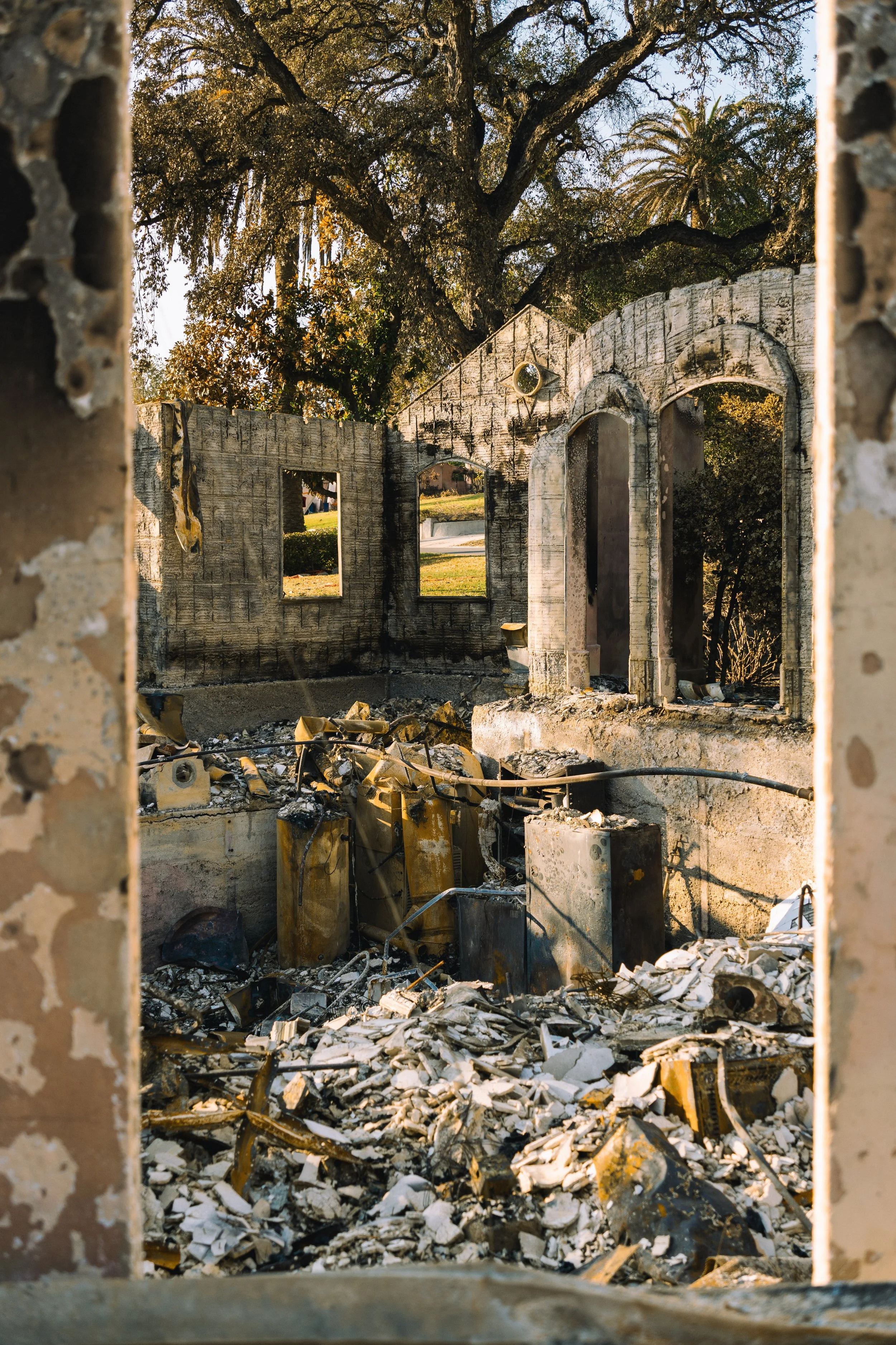 Ruined building viewed through a window frame with broken bricks and debris on the ground, partially covered by rubble, with trees in the background.