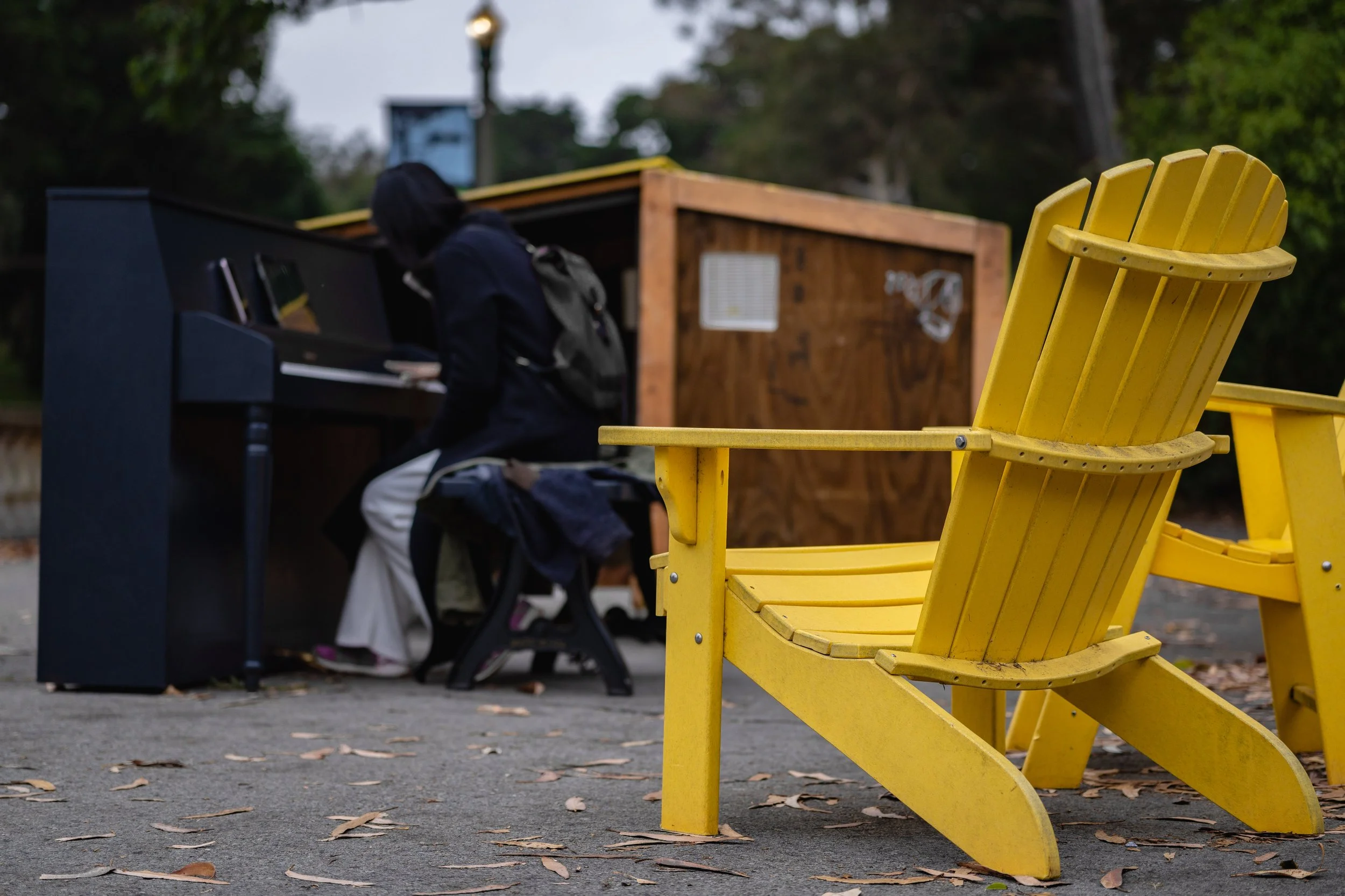 A yellow Adirondack chair in the foreground with a black music player and a backpack on a bench in the background, in an outdoor park setting during dusk.