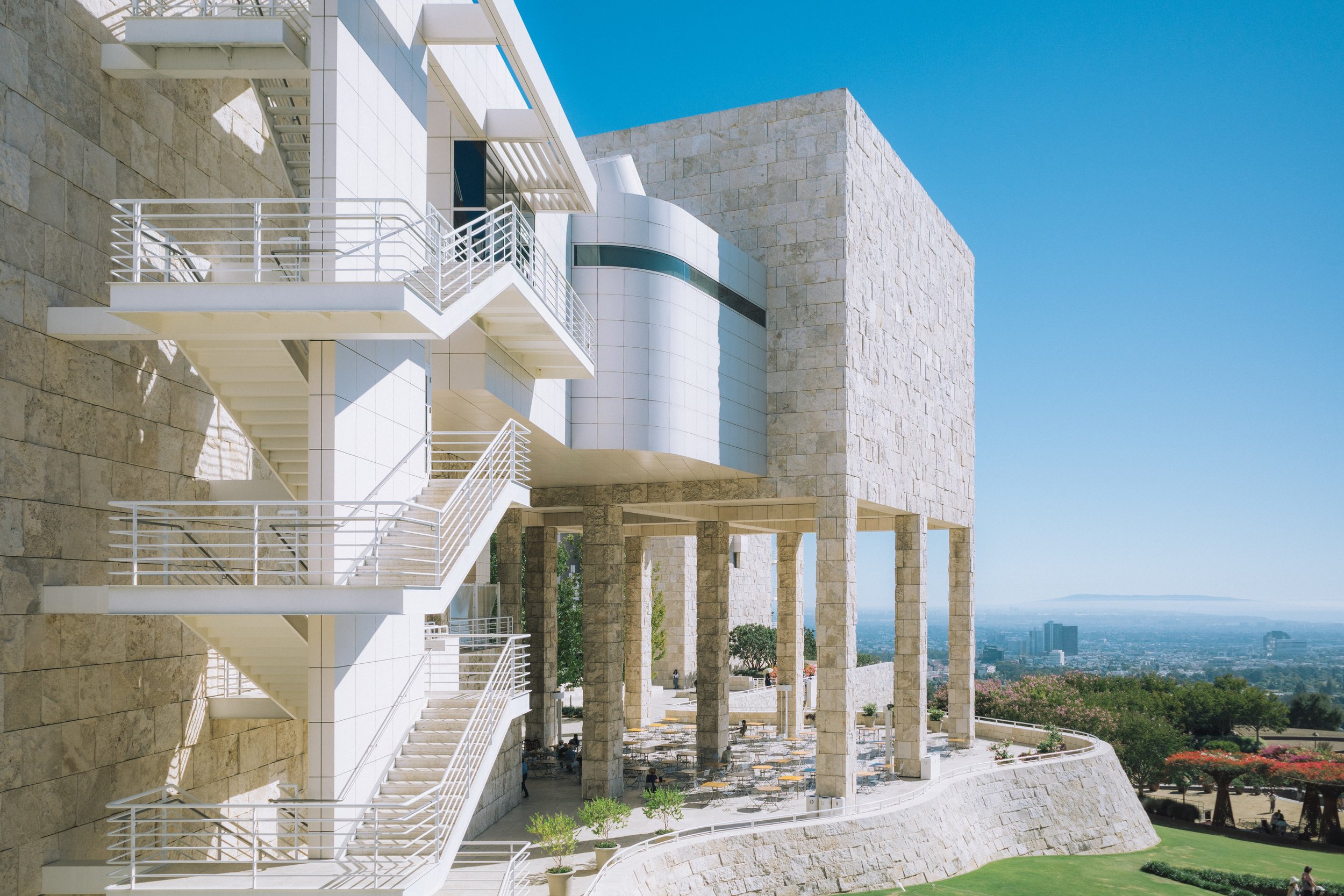 Modern building with white and beige stone facade, multiple levels with terraces and outdoor staircases, set against a clear blue sky with a cityscape in the distance.
