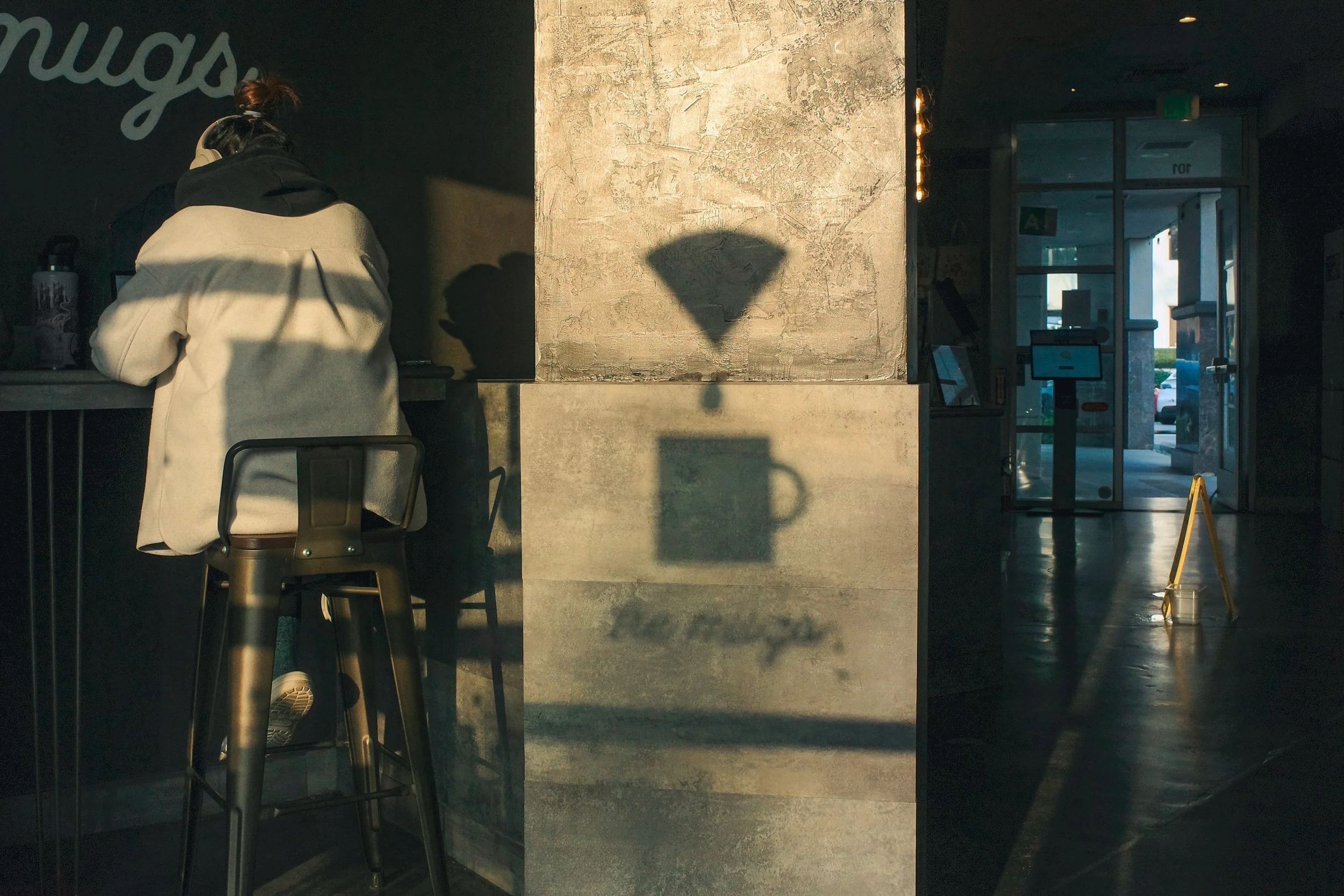Interior of a modern building with a person sitting at a counter. The person's shadow, cast by sunlight, is visible on a textured wall, and the entrance with glass doors is in the background.