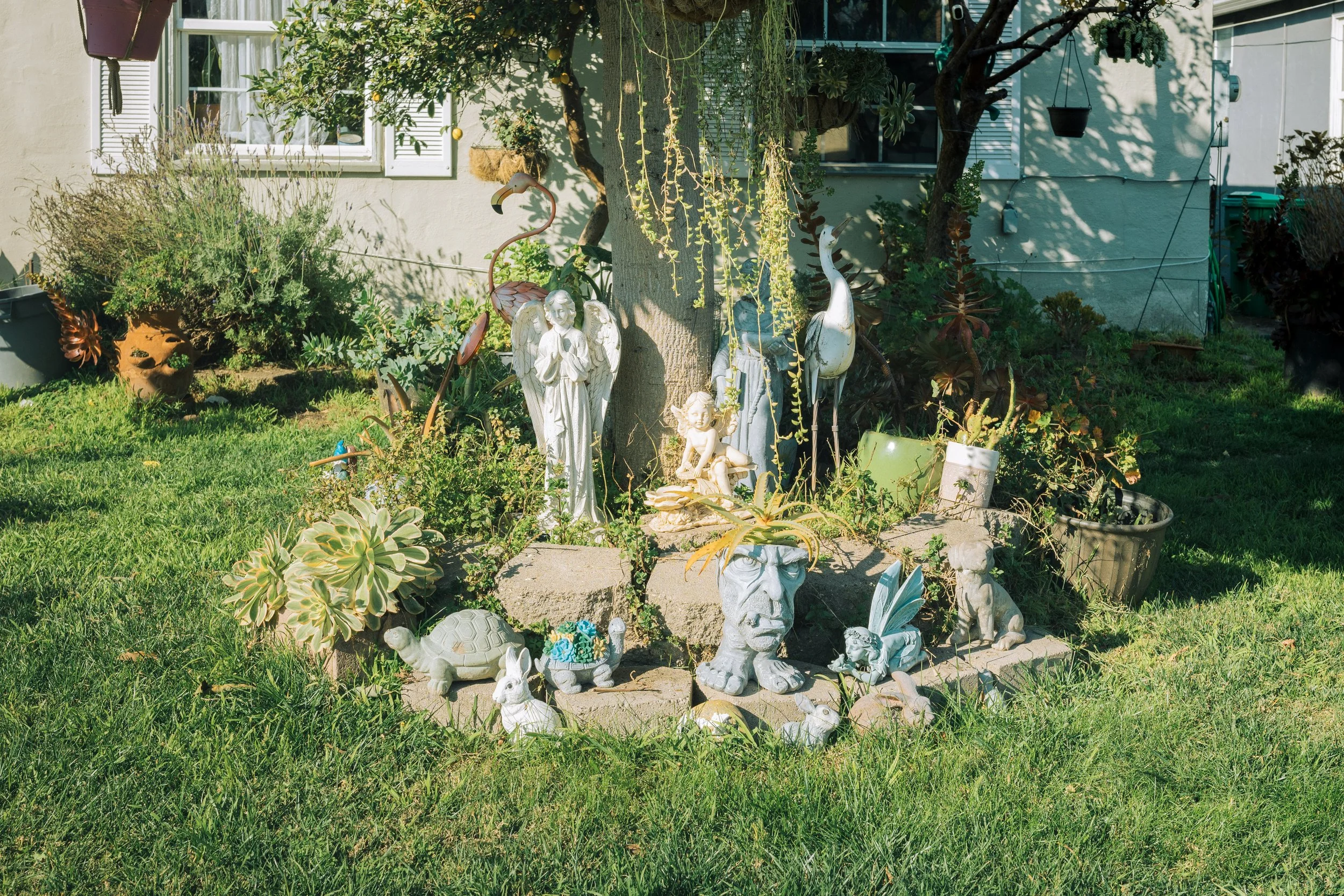 Garden yard with various decorative statues including angels, animals, and mythical creatures, surrounded by lush plants and grass, with houses and windows visible in the background.