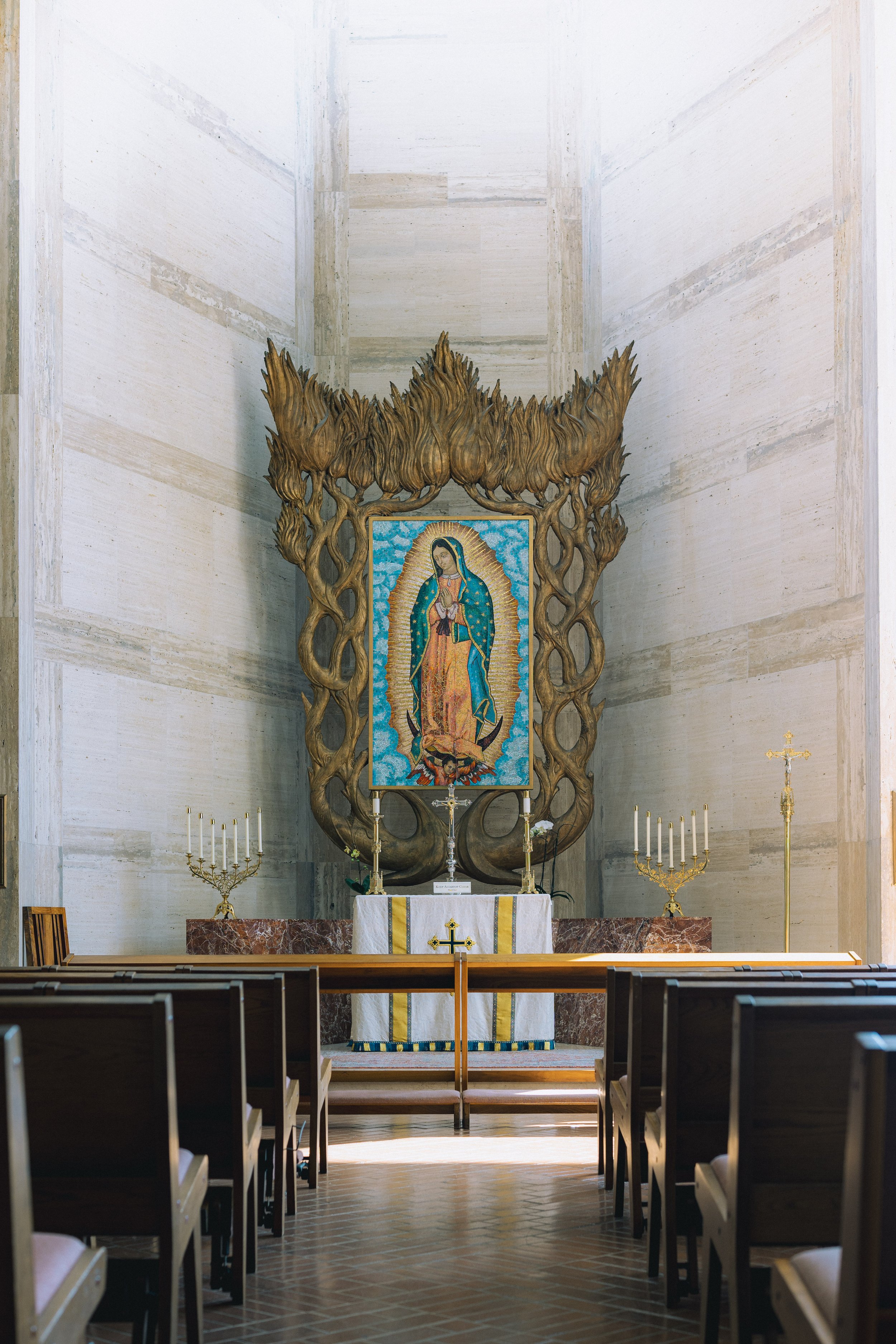 Interior of a church altar showcasing a large image of Our Lady of Guadalupe, framed by an ornate wooden structure, with lit candles and gold candelabras on either side.
