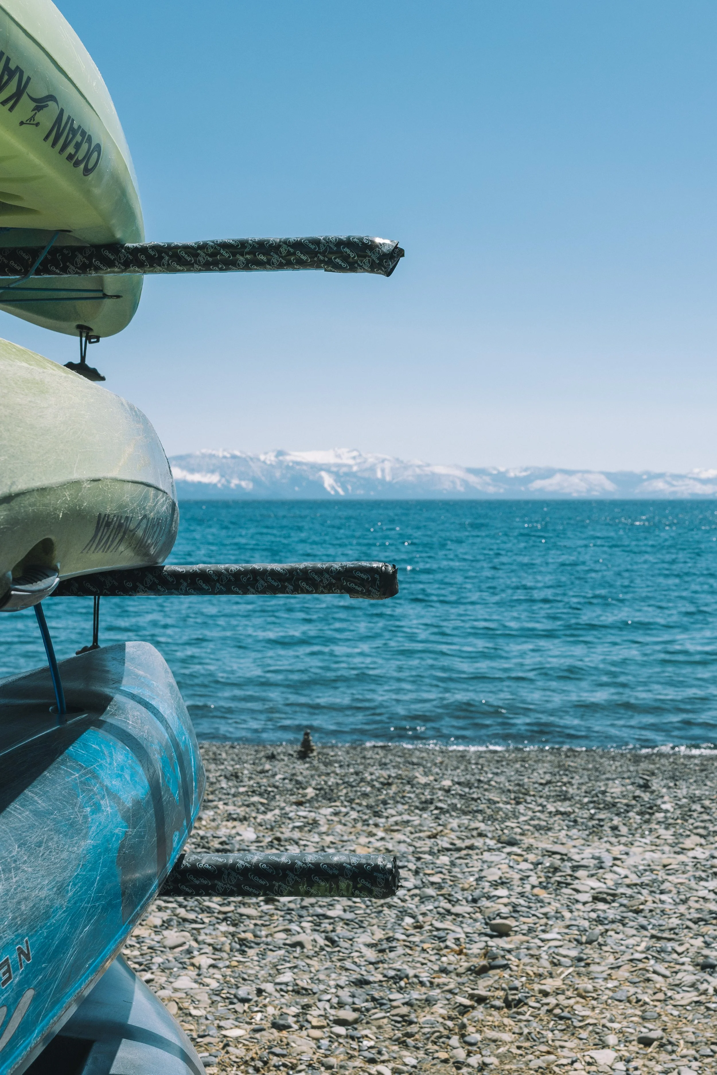 A line of colorful kayaks stacked on a rocky shoreline with calm water and snow-capped mountains in the distance under a clear blue sky.