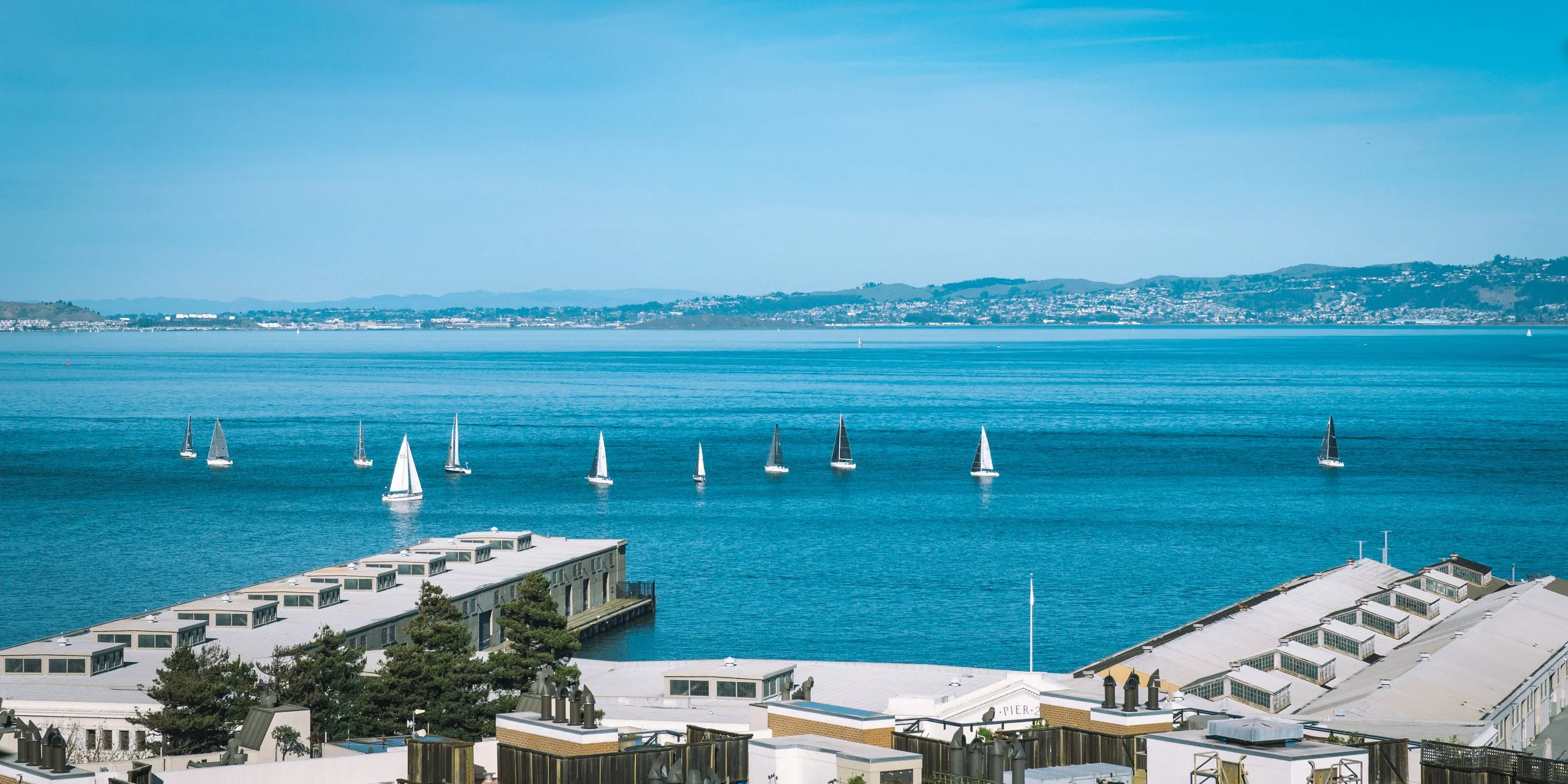 A view of the bay with sailboats on the water, a cityscape in the background, and modern rooftops and buildings in the foreground.