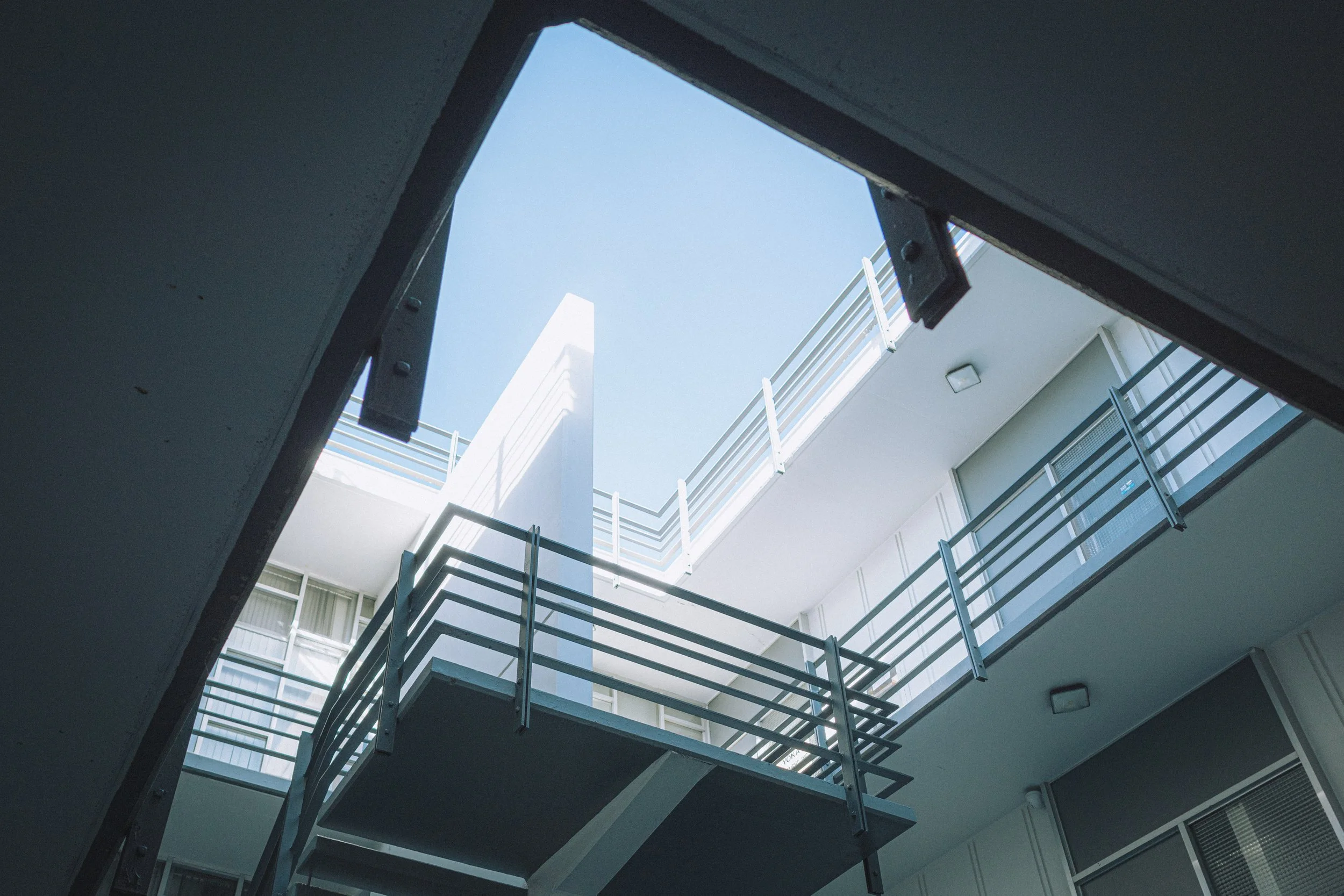 Looking up through the open ceiling of a modern multi-story building with white walls, metal railings, and balconies, under a clear blue sky.