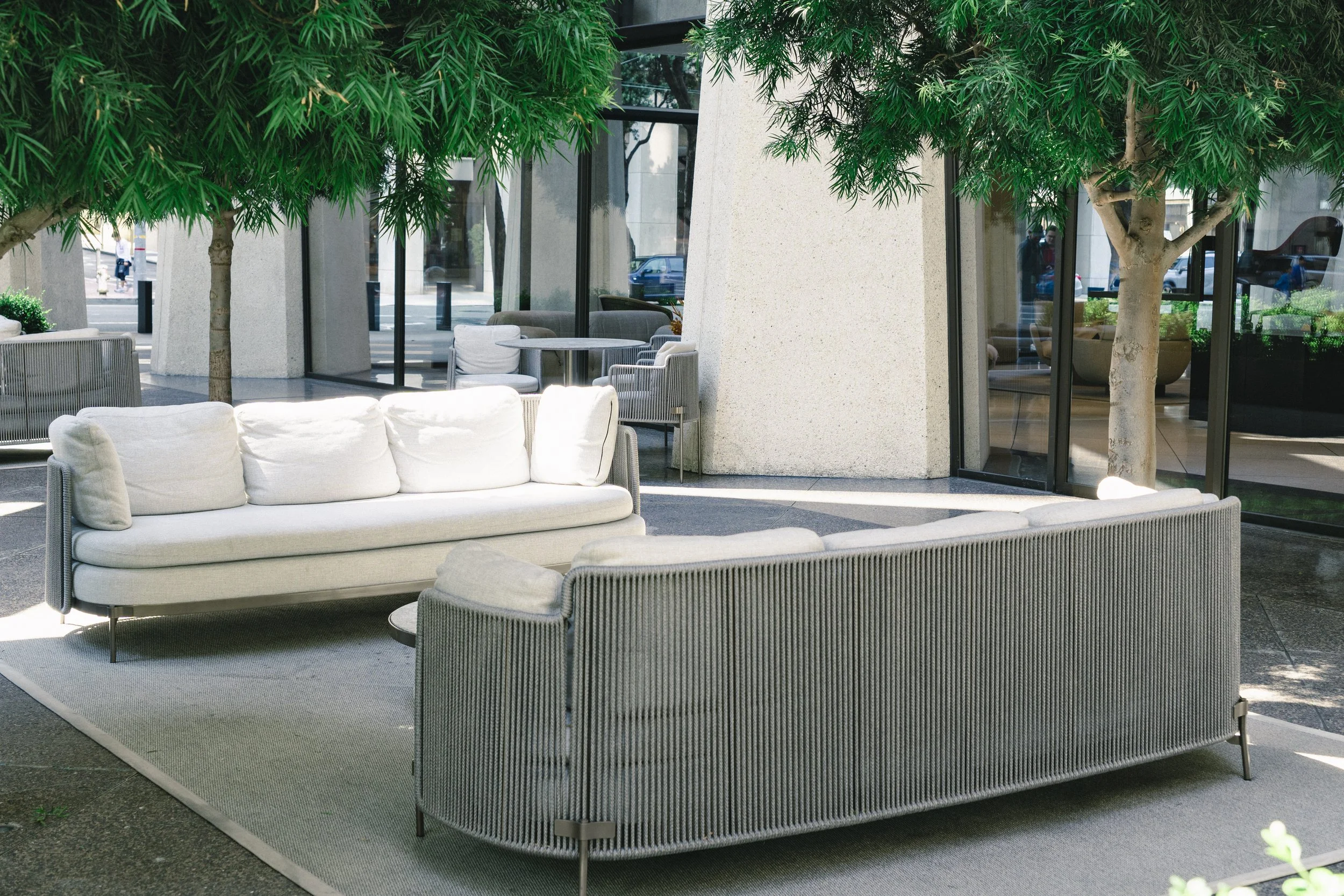 Modern outdoor seating area with white cushioned sofas and gray armchairs on a gray rug, topped with leafy green trees, situated in front of a glass building with reflections of people and cars outside.