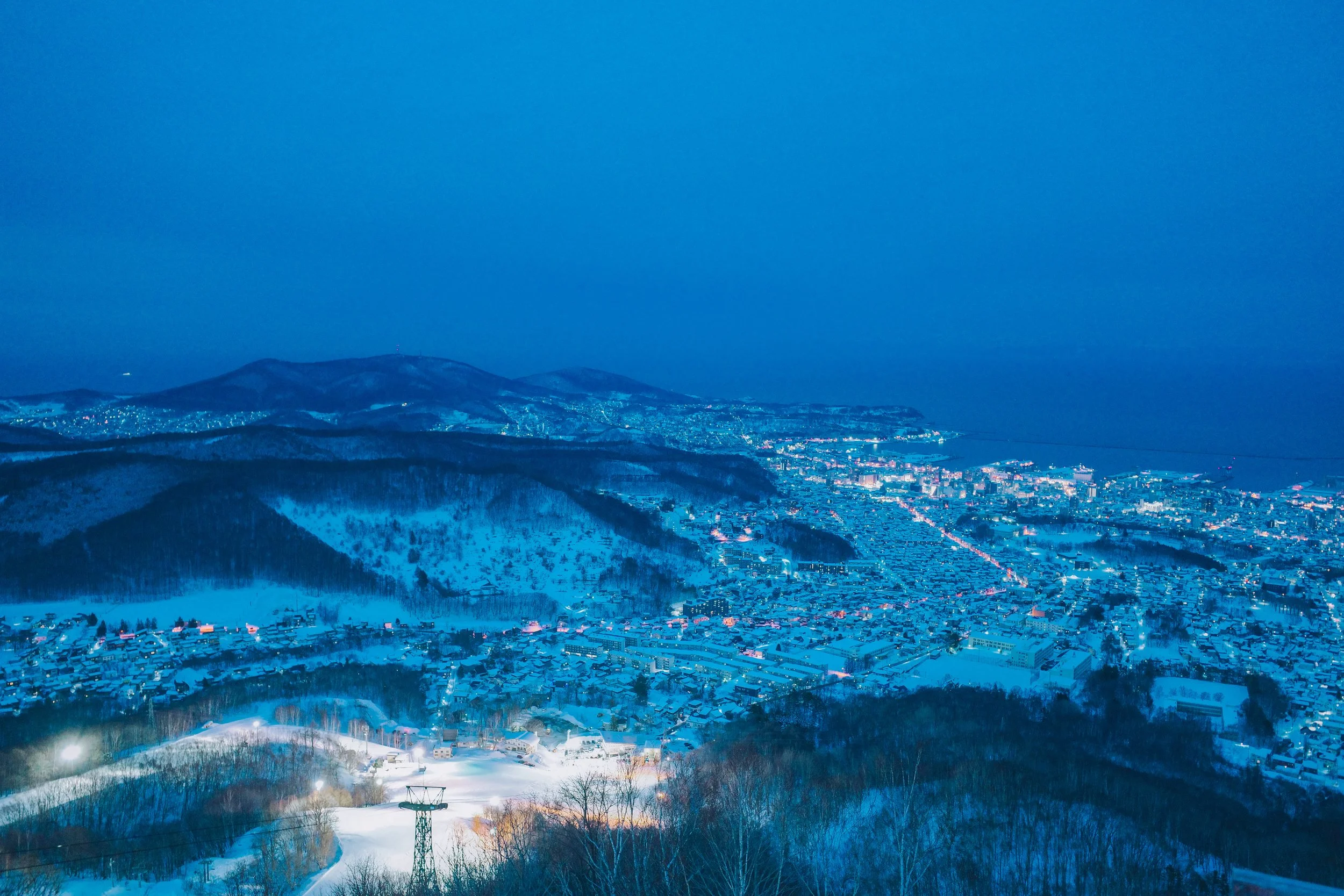 Nighttime view of a snow-covered town surrounded by mountains, with lights illuminating the area and a ski lift in the foreground.