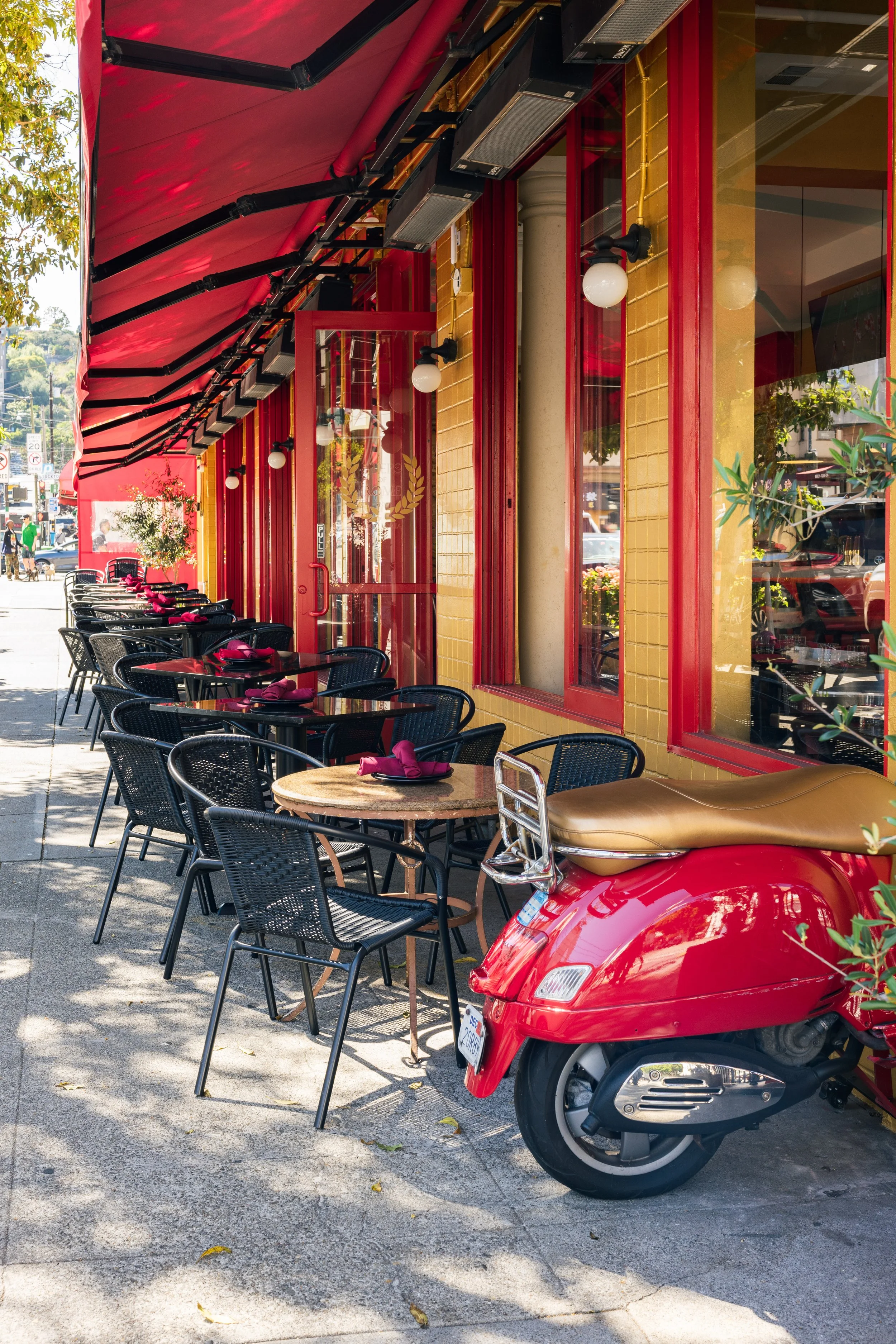 Outdoor restaurant seating with black metal tables and chairs, purple napkins, and a red car parked nearby on the sidewalk. The restaurant has large glass windows, red frames, and yellow brick walls, with a red awning overhead. Trees and street scene