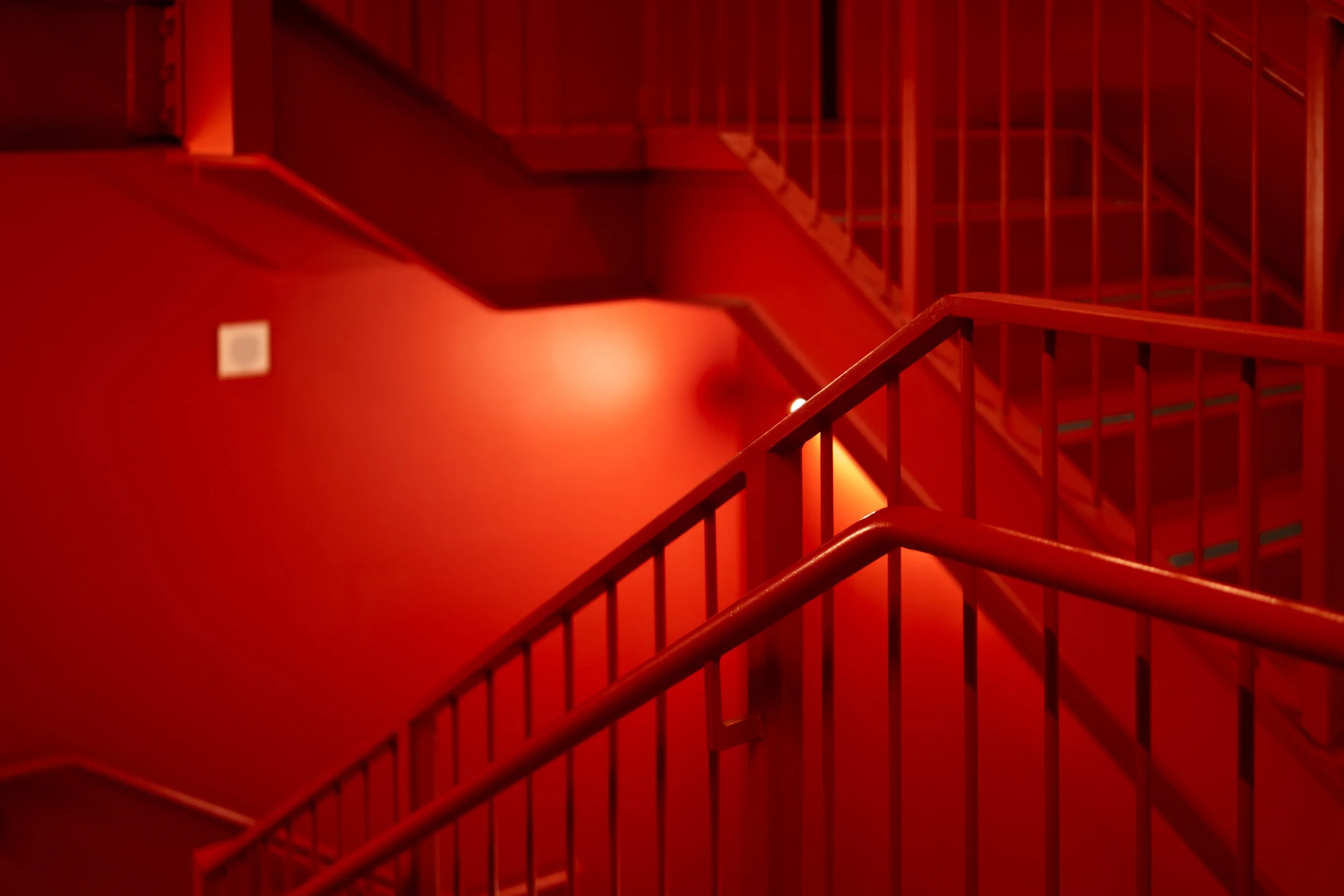 Red staircase with handrails and a wall with a small white vent, illuminated by warm red lighting.