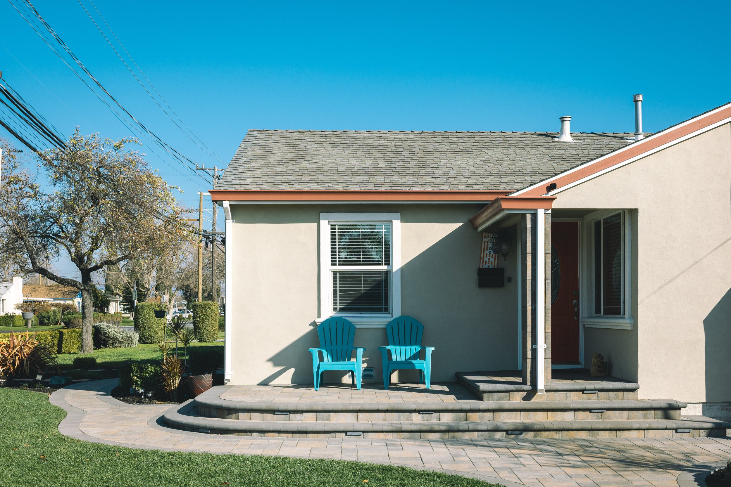 Front view of a house with a beige exterior, white trim, gray shingled roof, red door, and two blue chairs on a small patio with stone steps. There are trees, green lawn, and power lines in the background.