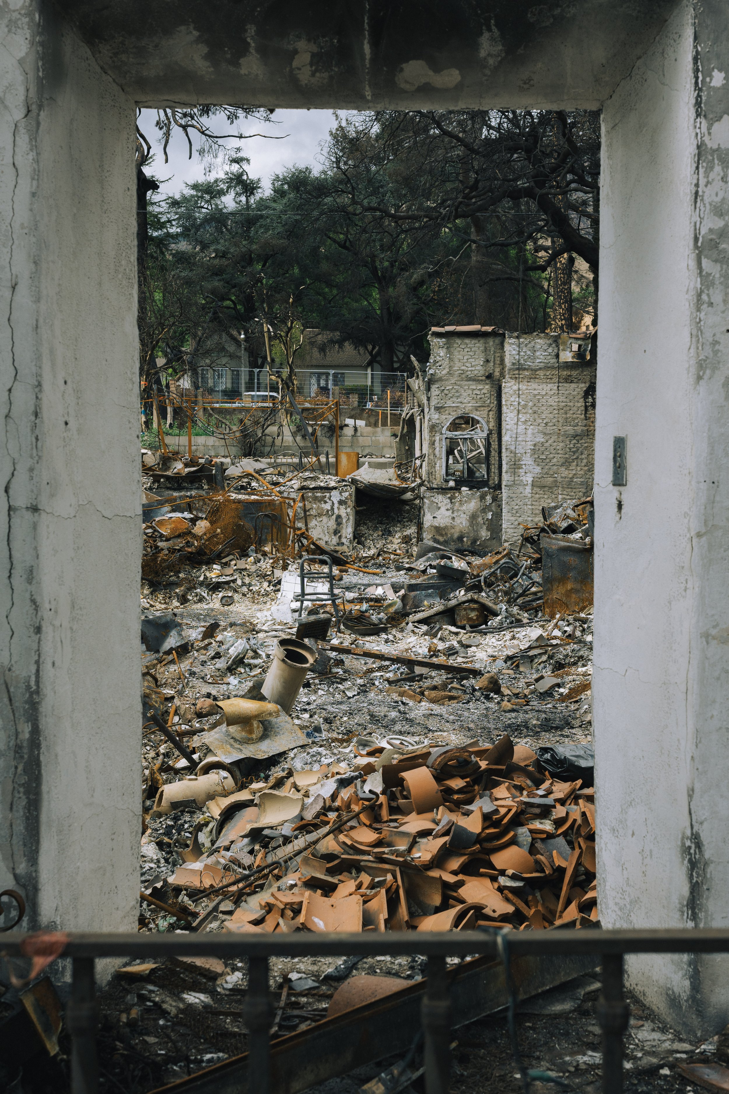 View of a burned house with debris and ruins seen through a window frame, with blackened walls and charred remains of the structure and furniture.