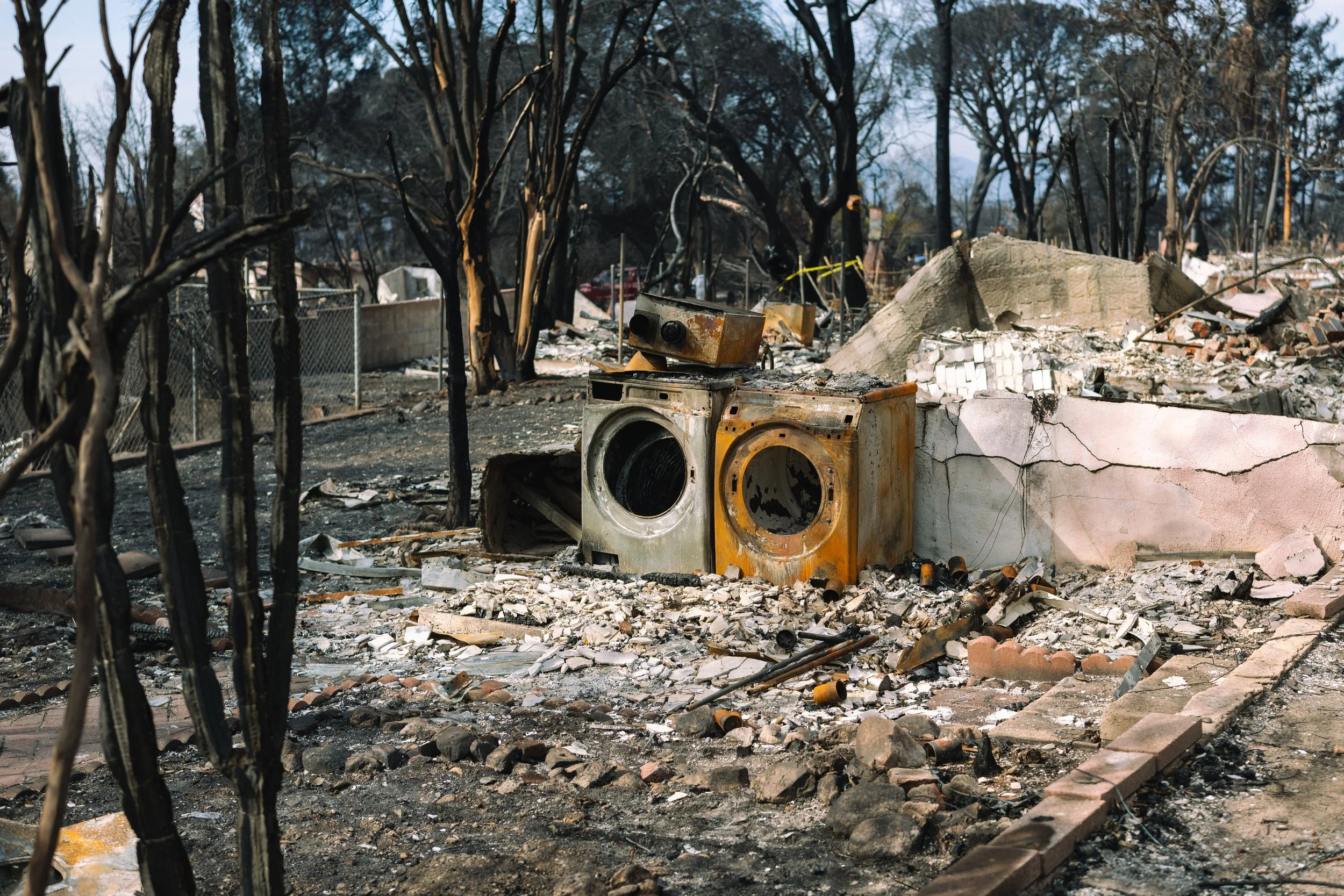 Burned and destroyed outdoor area with damaged appliances, charred trees, and debris from fire.