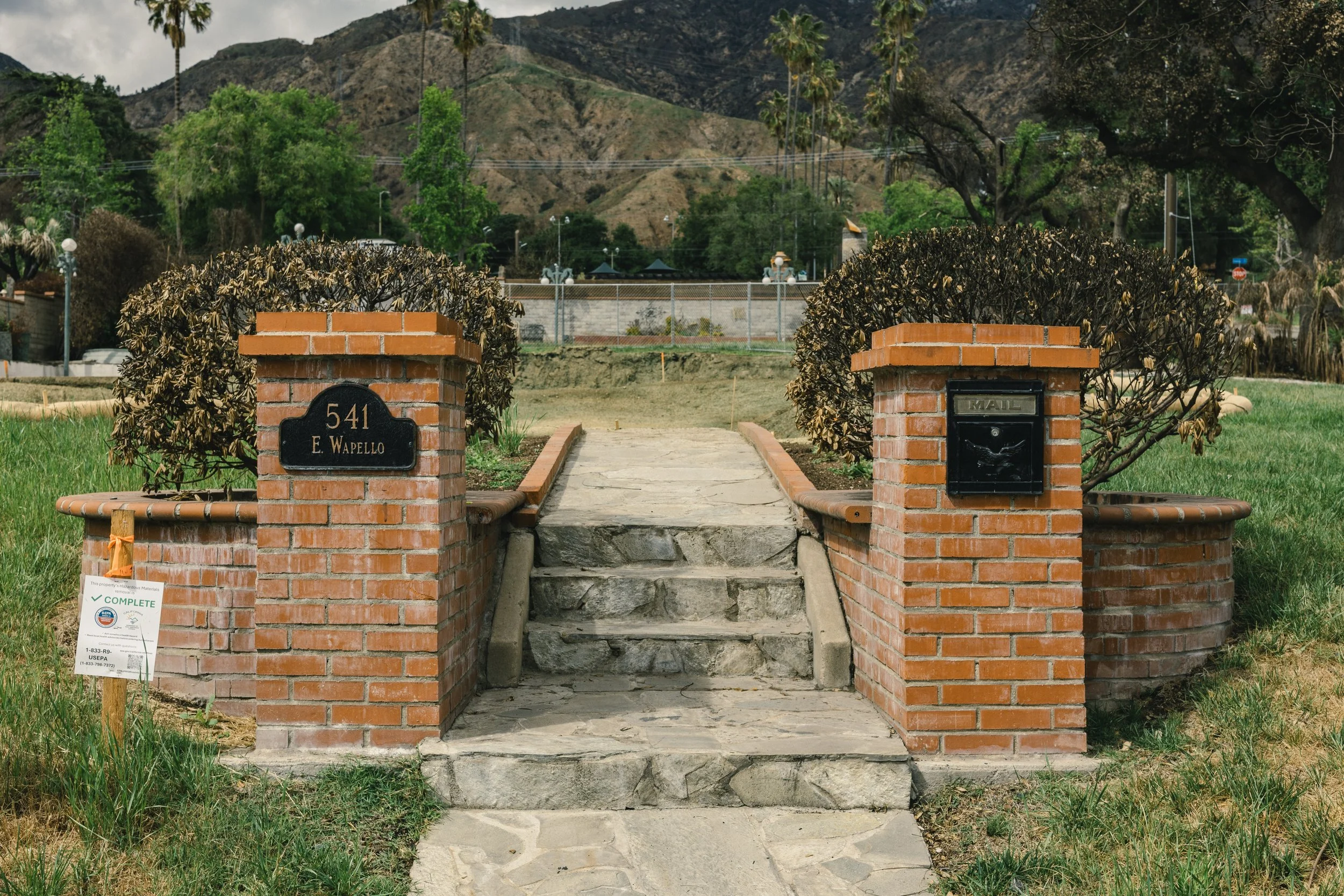 Front yard with brick steps leading to a sidewalk, flanked by brick and bush planters, in front of a house with a yard enclosed by a fence, hills in the background, and a cloudy sky.