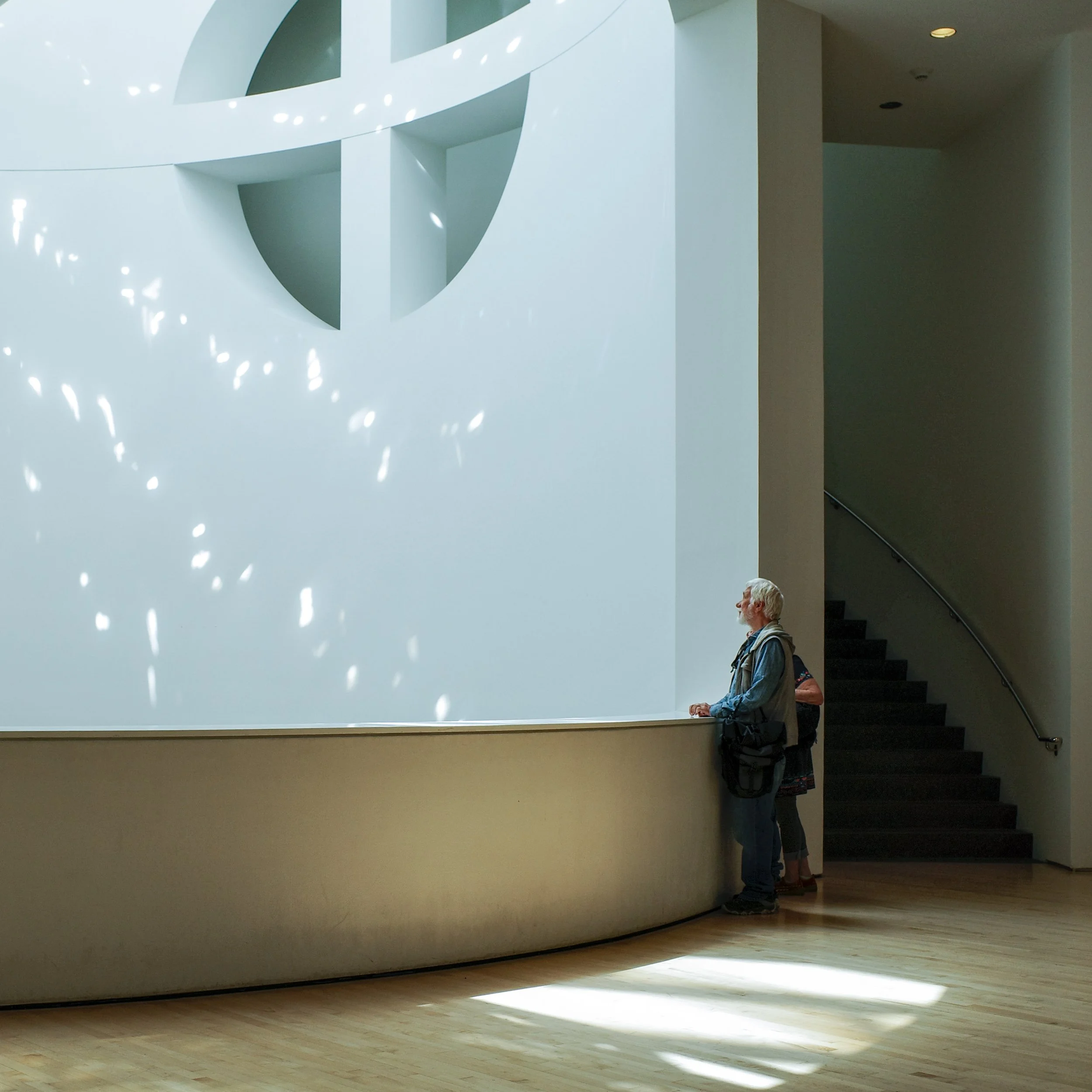 People standing in line inside a modern building with white walls, wooden flooring, and a staircase. Light streams through windows creating patterns on the wall.