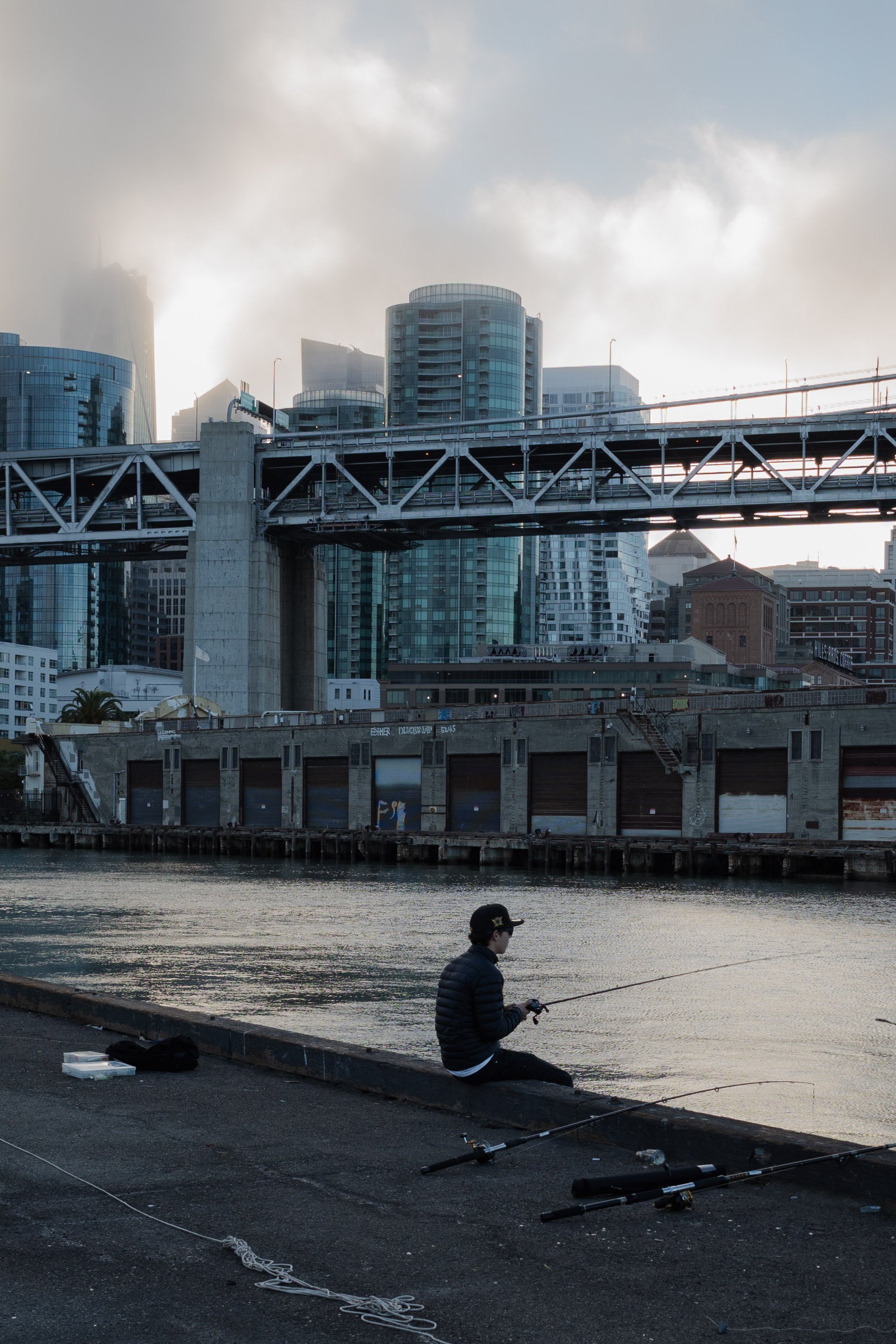 A person fishing by a river with an urban cityscape in the background, featuring high-rise buildings, a bridge, and a cloudy sky.