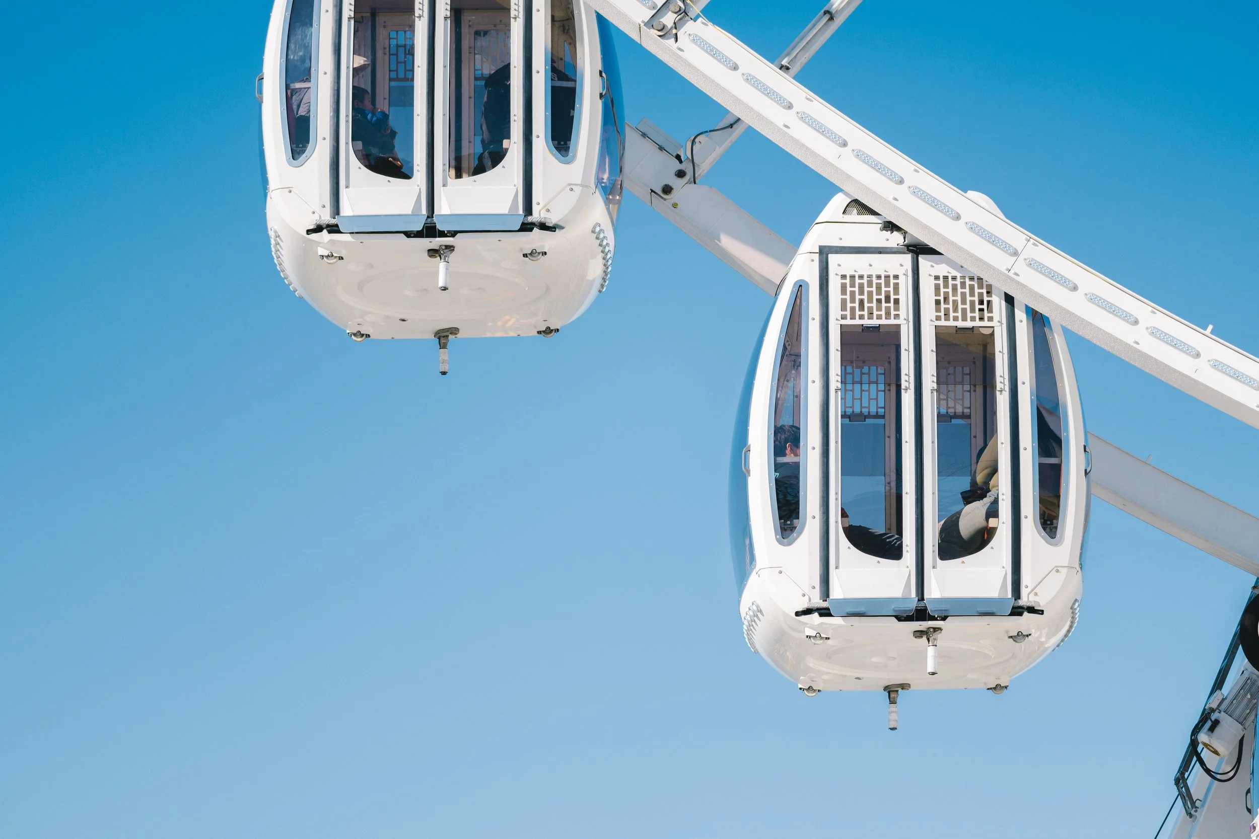 Close-up of a white Ferris wheel against a clear blue sky, with enclosed gondolas and visible passengers inside.