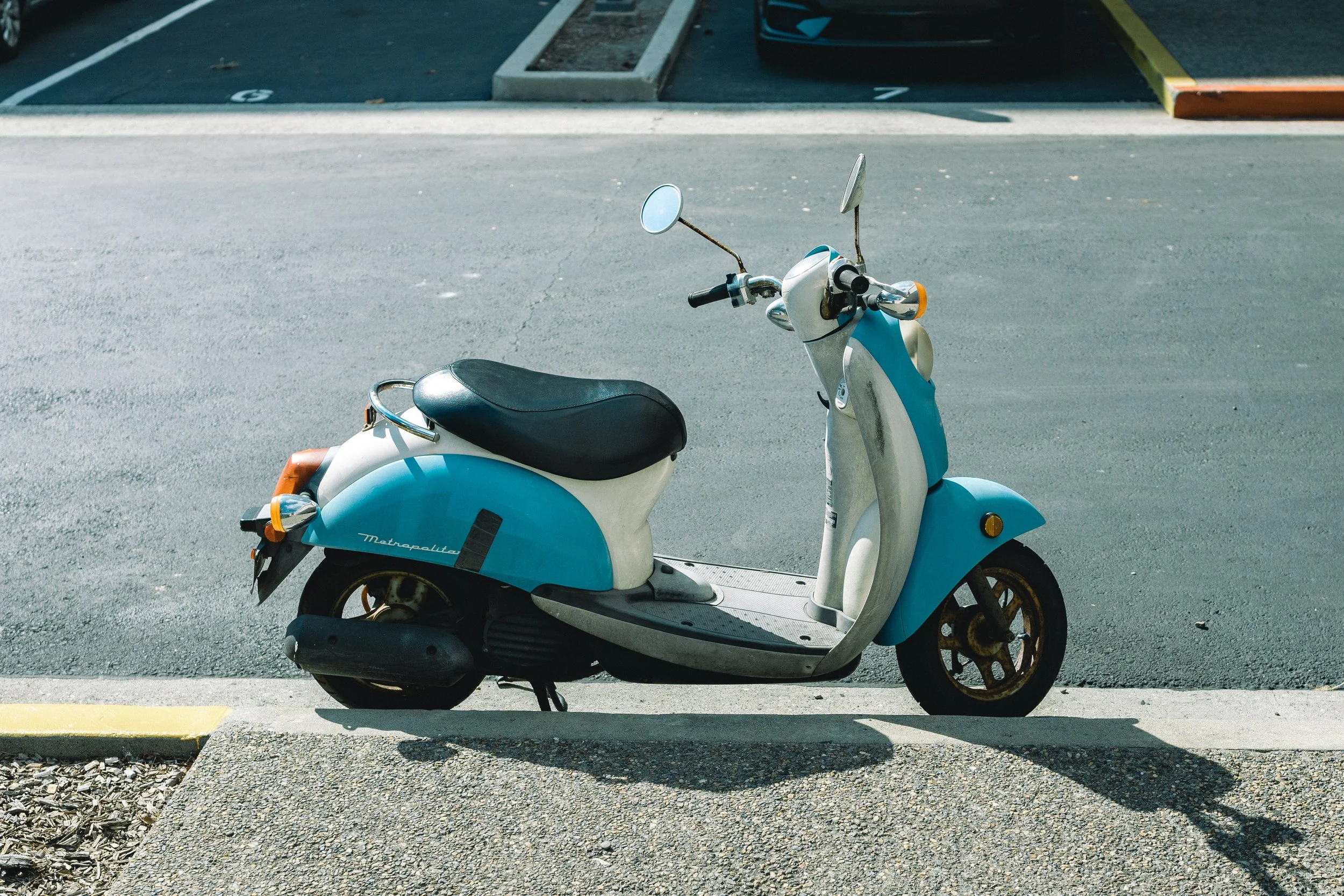 A blue and white Vespa scooter parked in a parking lot next to a curb and a parking space marked with the number 5. The scooter has a black seat and round mirrors.
