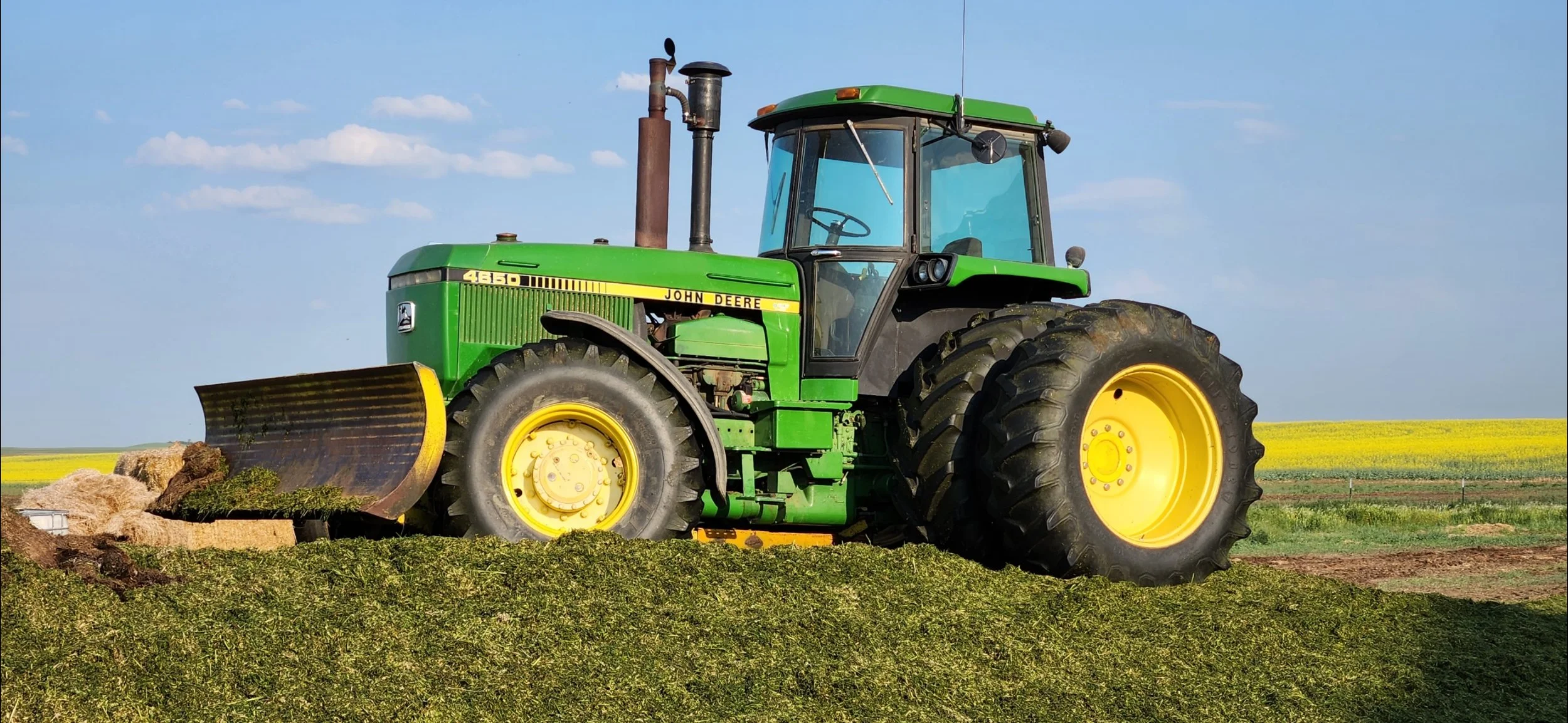 Green John Deere tractor with a snowplow attached, parked on a farm field under a blue sky with some clouds.