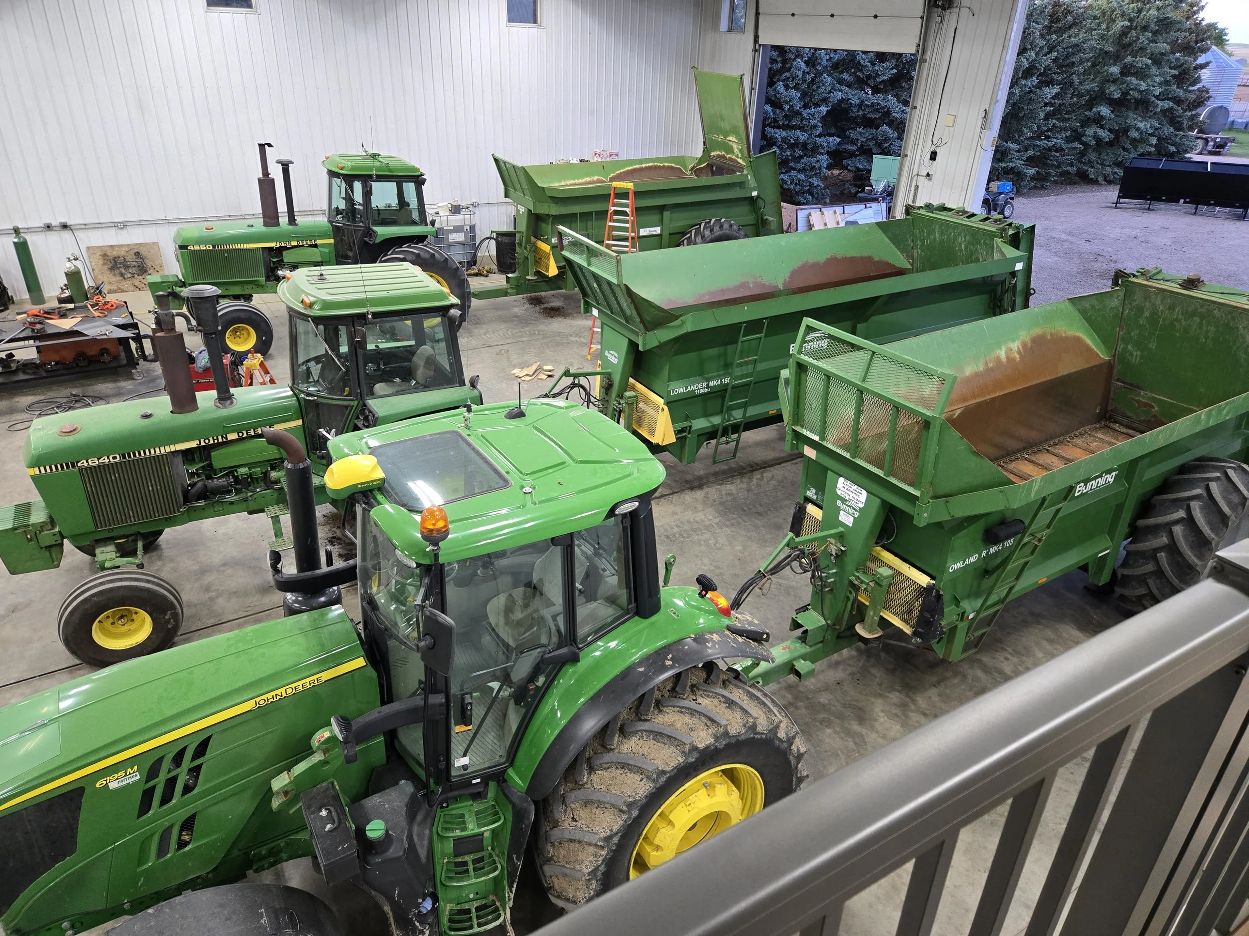 Multiple green John Deere tractors and trailers inside a shed with some tools and equipment nearby.