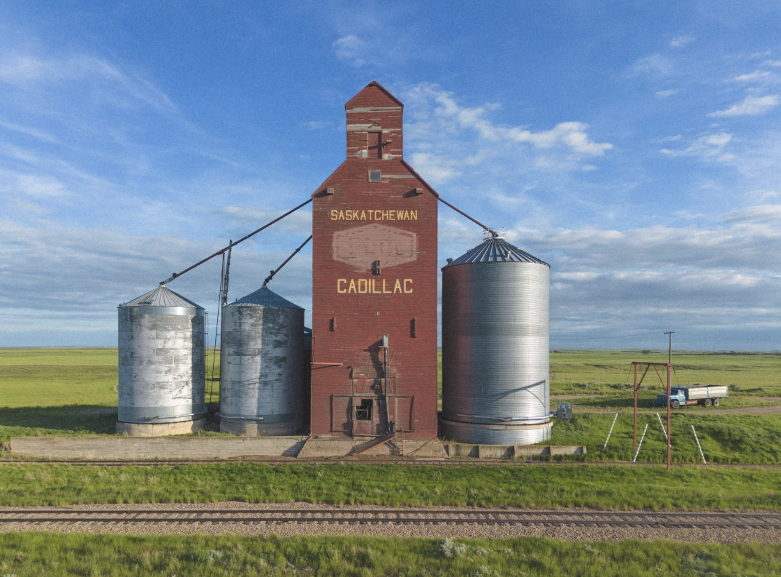 A red grain elevator with the words 'Saskatchewan Cadillac' on it, flanked by two silver silos, in a rural area under a partly cloudy blue sky, with a train track and a blue truck with a trailer in the background.