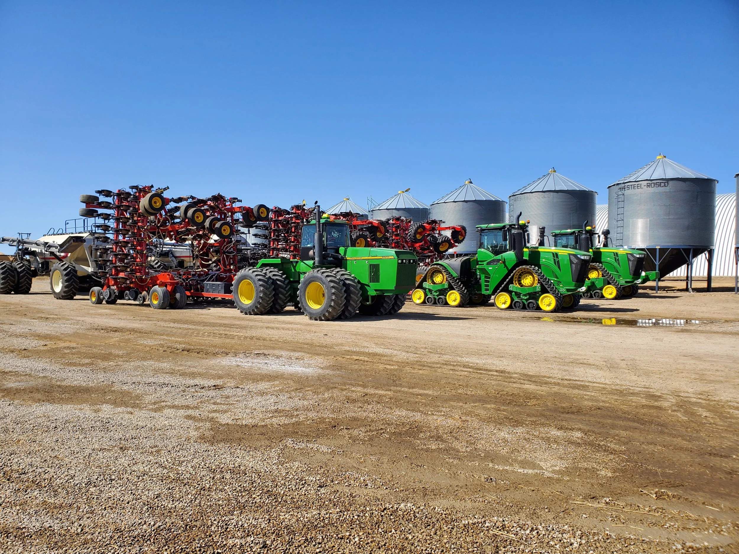 Farm equipment and large grain silos on a farm under a clear blue sky.