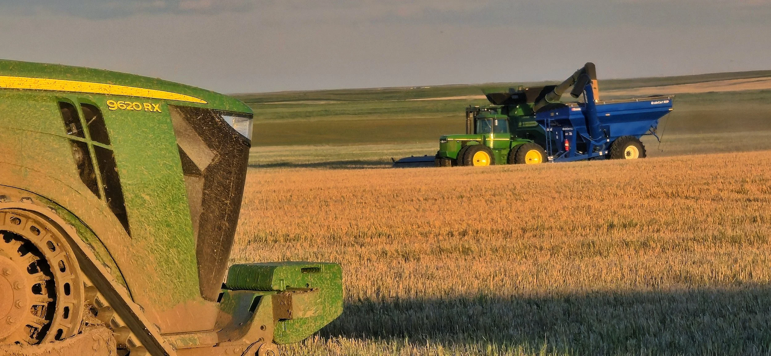 Close-up of a green John Deere 9620RX tractor in a wheat field with two blue and green harvesters in the distance under a partly cloudy sky.