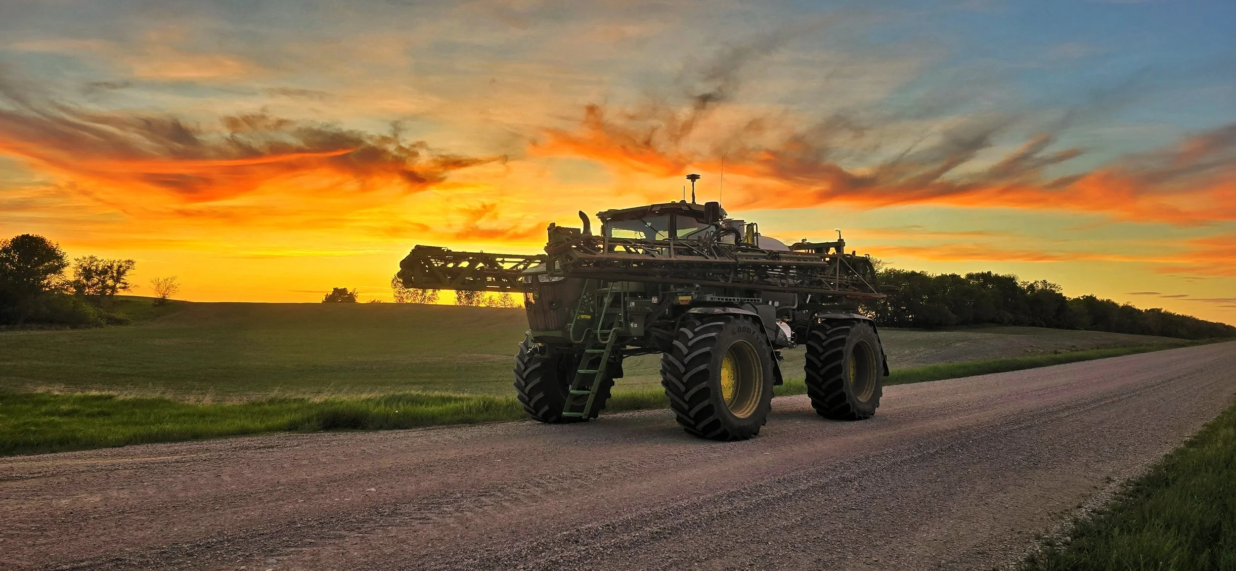 A large piece of agricultural equipment, specifically a sprayer, parked on a rural road during sunset with vibrant orange, yellow, and blue sky and trees in the background.
