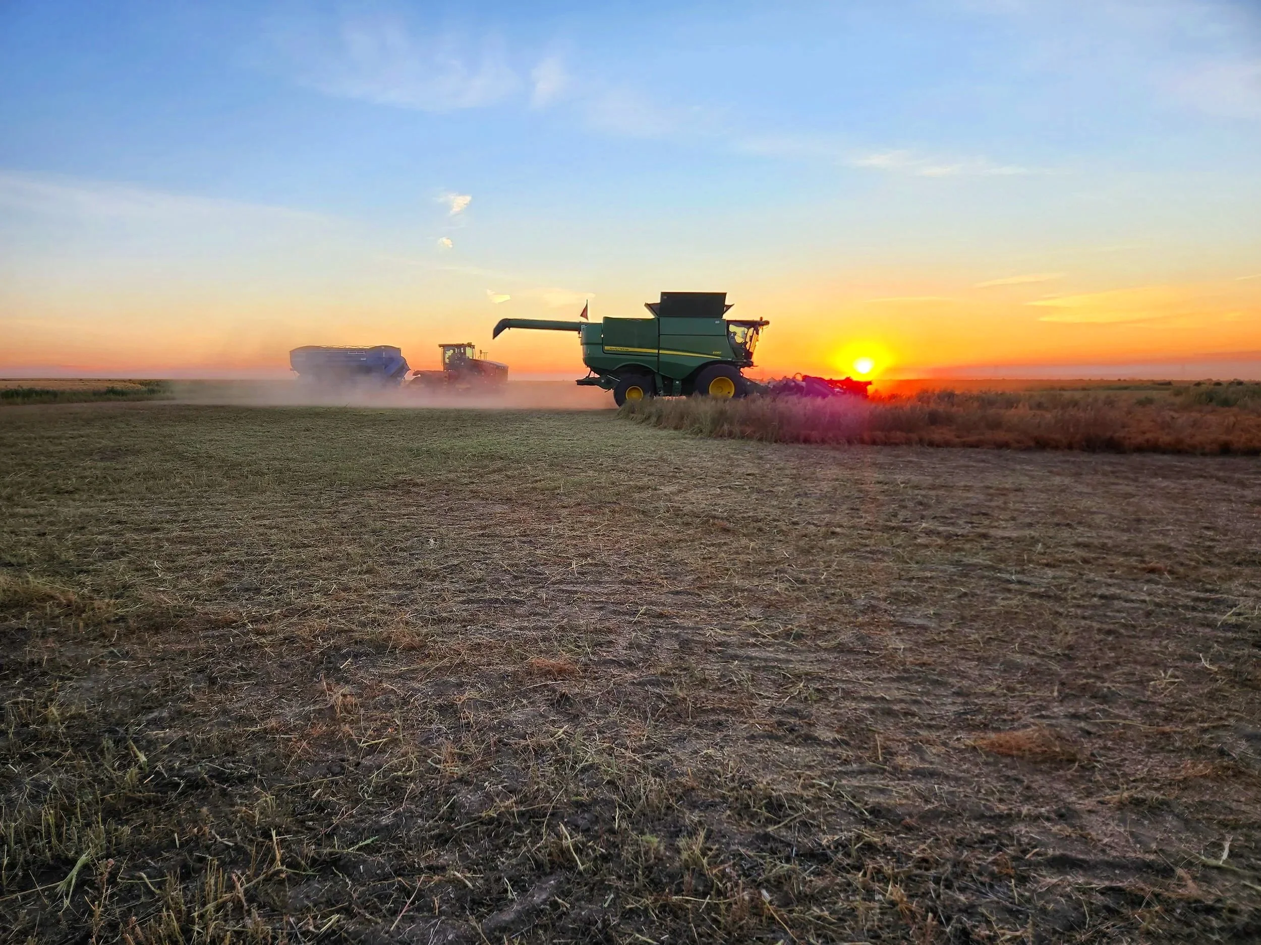 Farm equipment working on a field during sunrise, with a combine harvester and tractor creating dust, under a reddish-orange sky.
