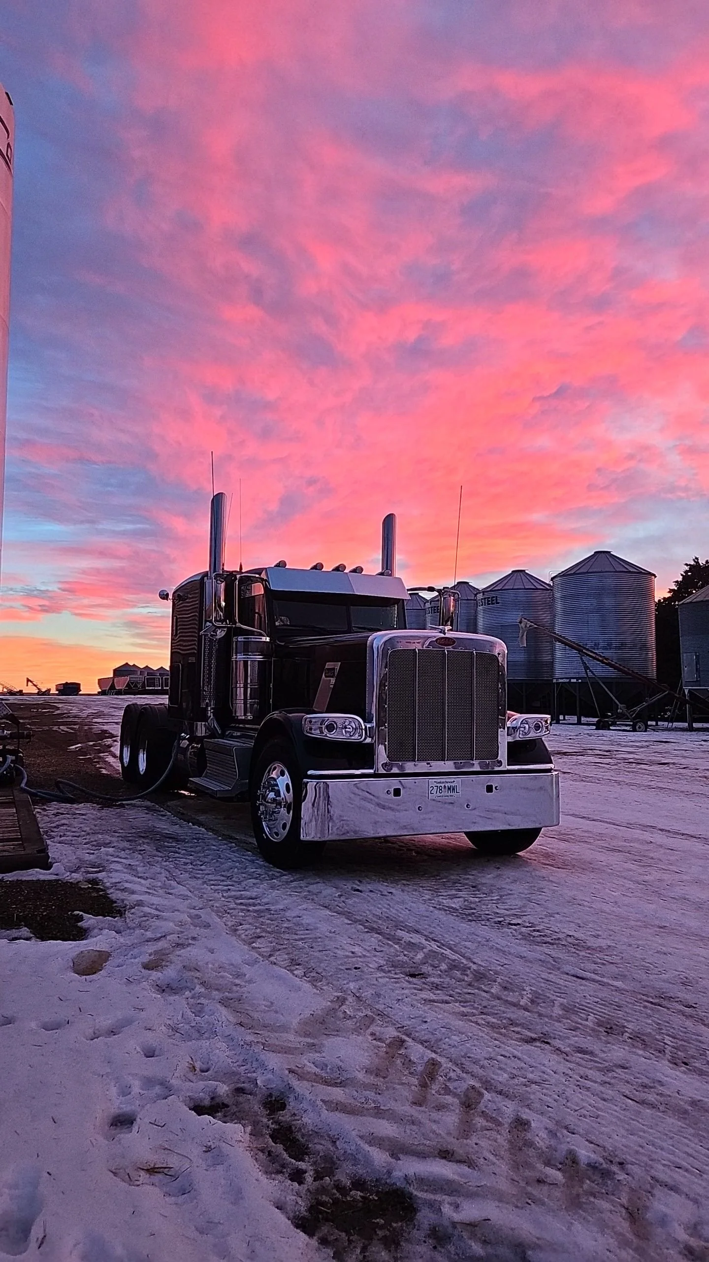 A black semi-truck parked on a snowy farm at sunset, with pink and purple clouds in the sky and metal grain silos in the background.