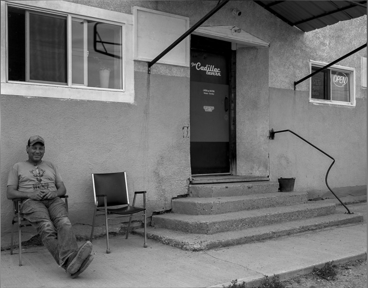 A man sitting on a chair outside a building with a couple of chairs next to him, the building has steps leading up to a door with an 'Open' sign in a window, and another window above the door.