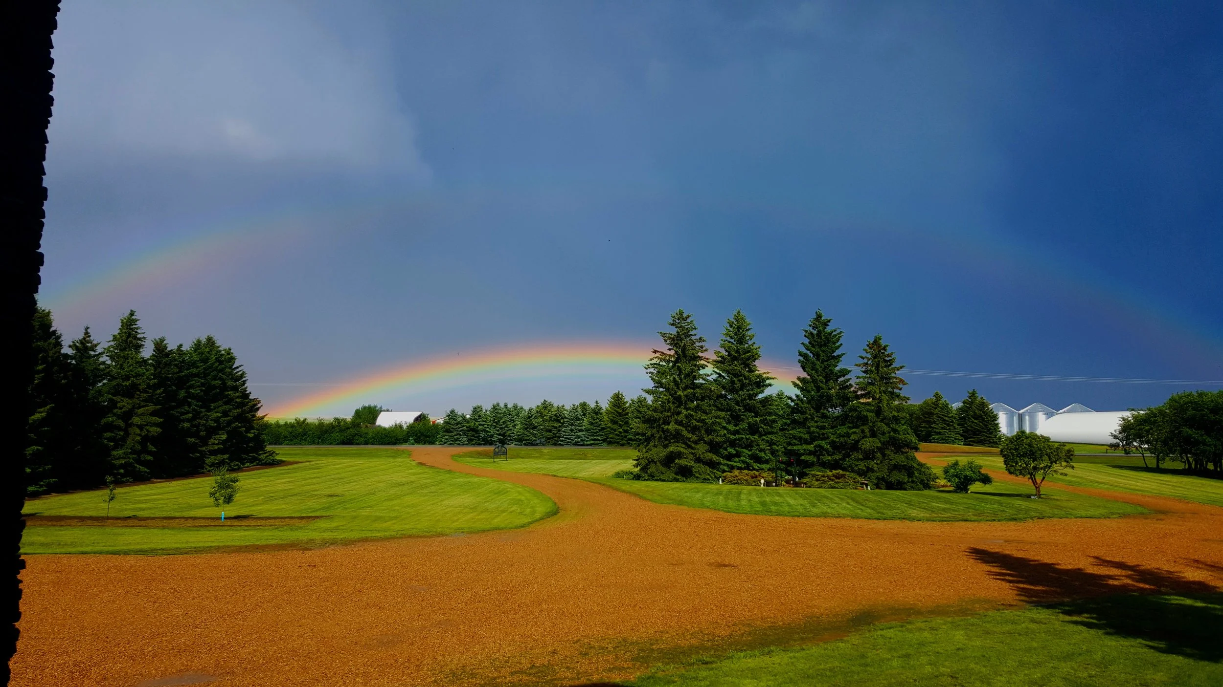 A scene of a farm landscape with green grass, trees, farm buildings, a dirt path, a rainbow in the sky, and storm clouds overhead.