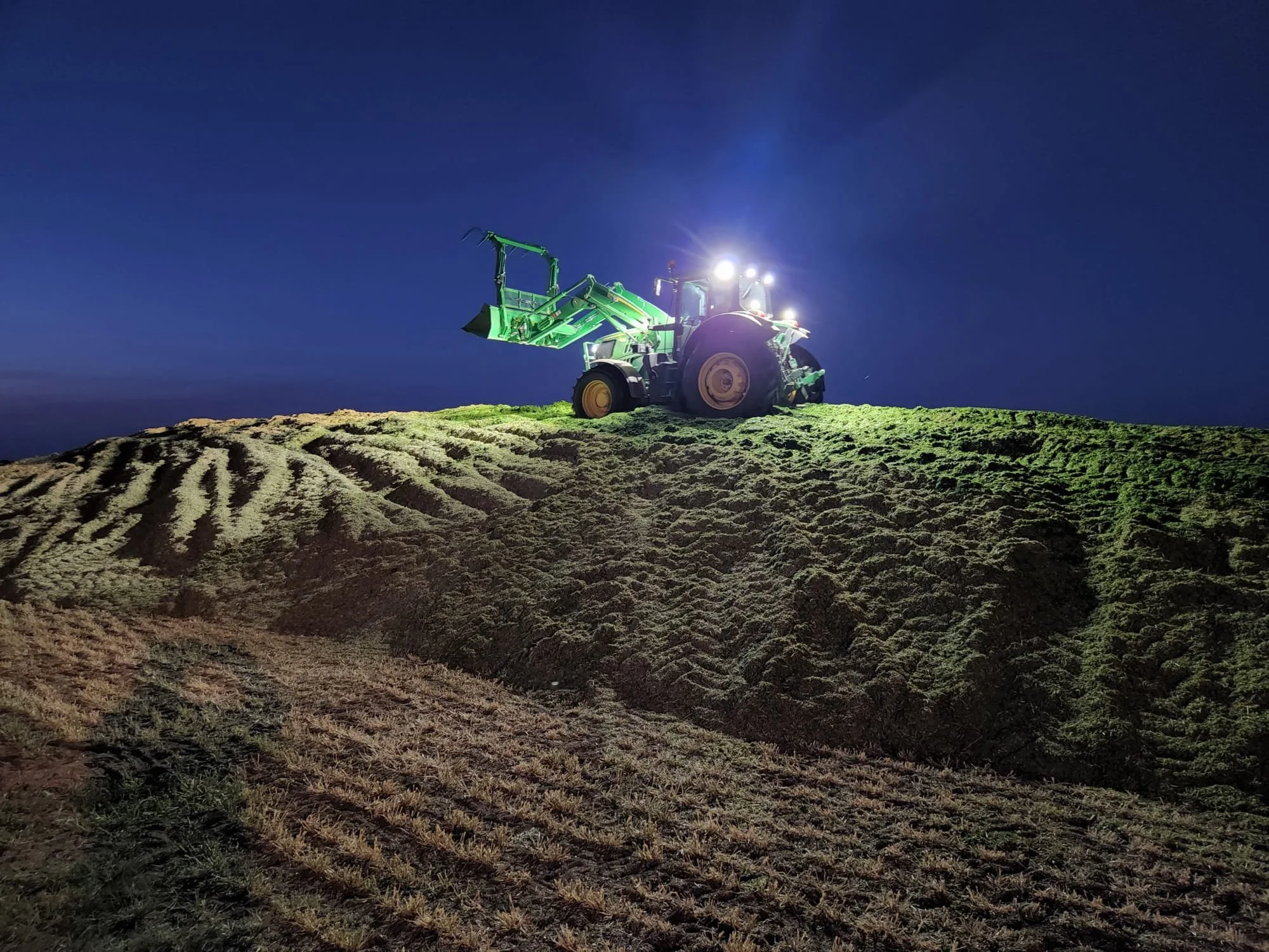 A green tractor working on a hillside at night, illuminated by bright lights against a dark blue sky.
