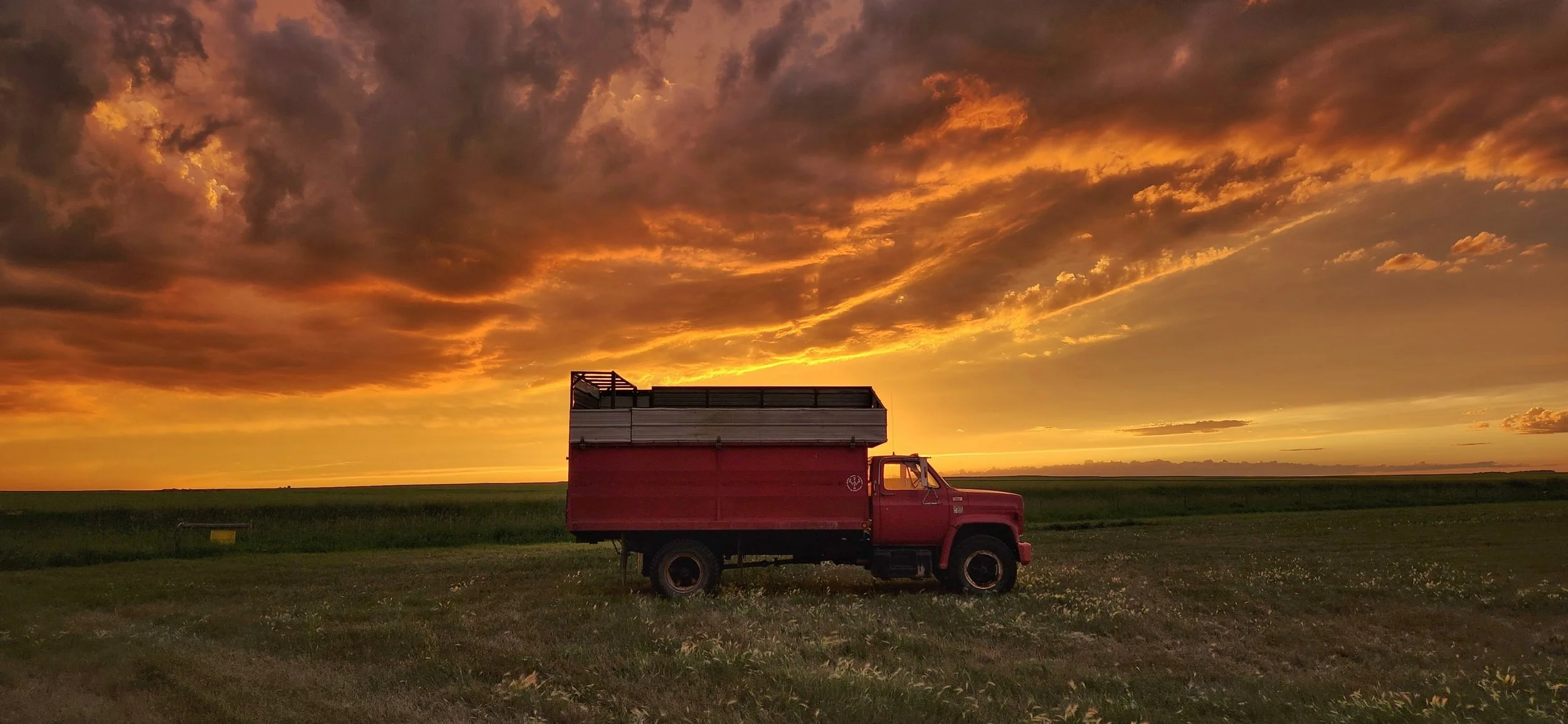 A red truck parked in a field at sunset with a sky filled with orange and dark clouds.