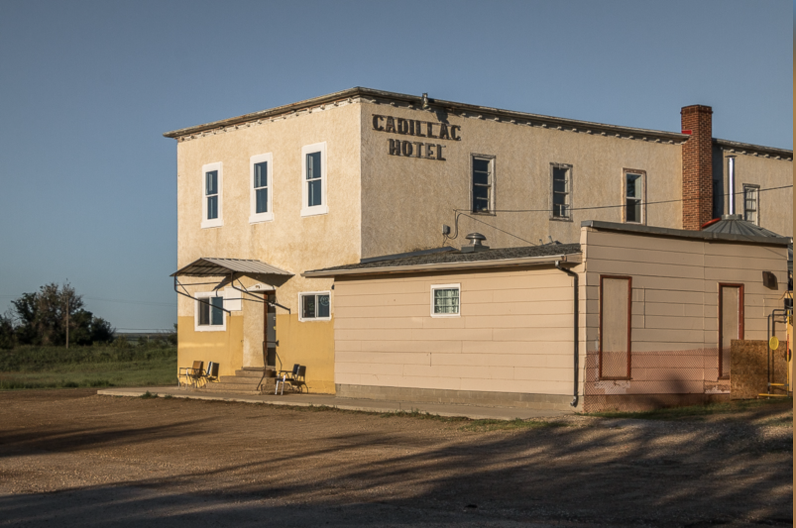 A two-story beige and yellow building with a sign that reads Cadillac Hotel, with a small entrance porch, several chairs outside, and a clear blue sky in the background.