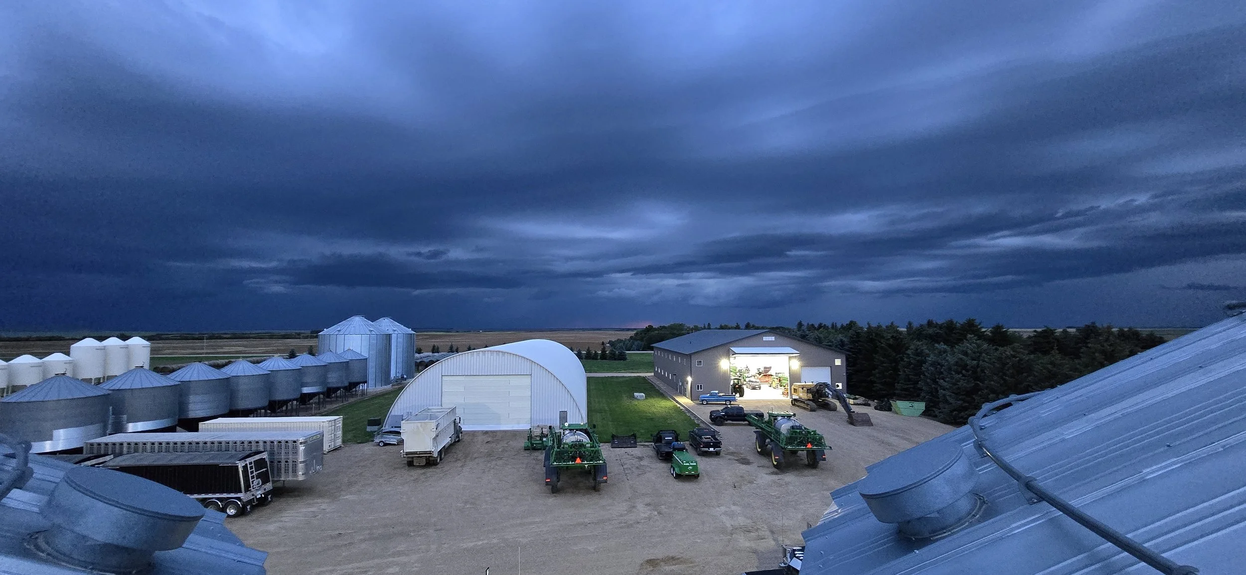View of a farm with silos and a barn under a dark, cloudy sky at dusk or dawn, with farm vehicles and equipment visible.