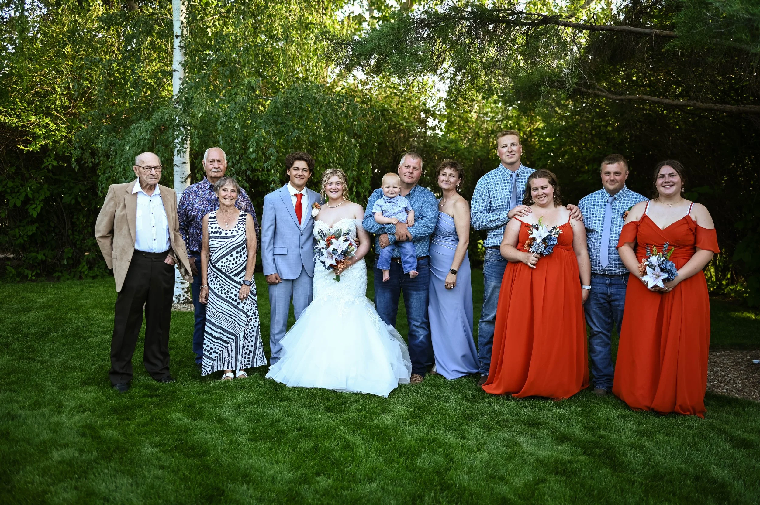 A large group of family members standing outdoors on a grassy area beneath trees during a wedding celebration. The bride and groom are in the center, with the bride wearing a white lace wedding dress and holding a bouquet, and the groom in a light blue suit with a red tie. The family members are dressed in various colorful and formal attire, some holding flowers, smiling and posing together.