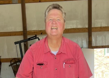 A woman with short gray hair wearing a pink shirt, smiling, in an indoor setting with wooden beams and a metal structure in the background.
