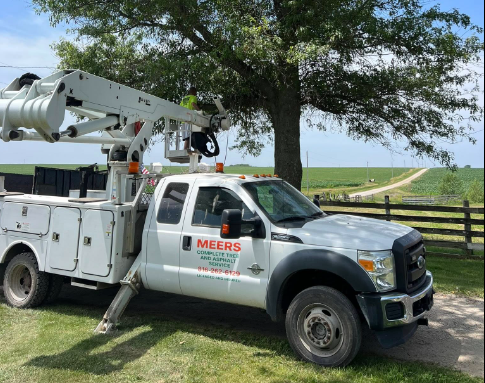 Utility truck with a bucket lift parked on grass near a tree, with a rural background of open fields and a dirt road.