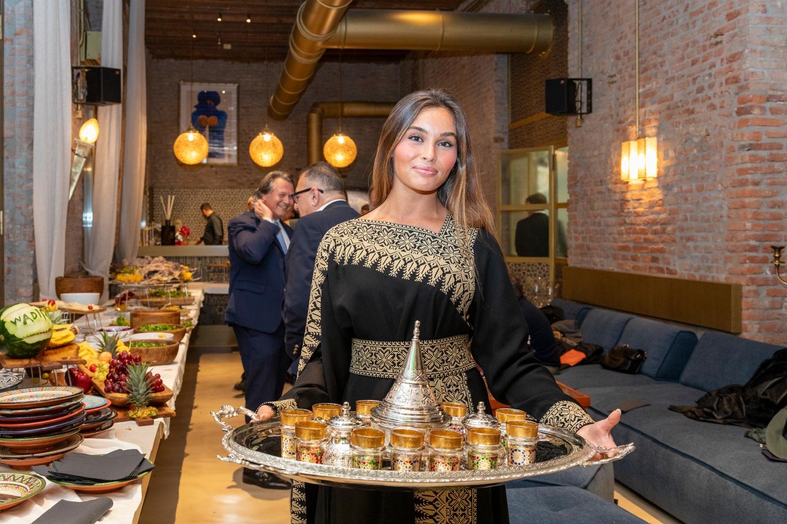 Una giovane donna che sorride tiene in mano un vassoio con coppe di vetro e una teiera d'argento, in un ristorante con pareti di mattoni e luci calde.