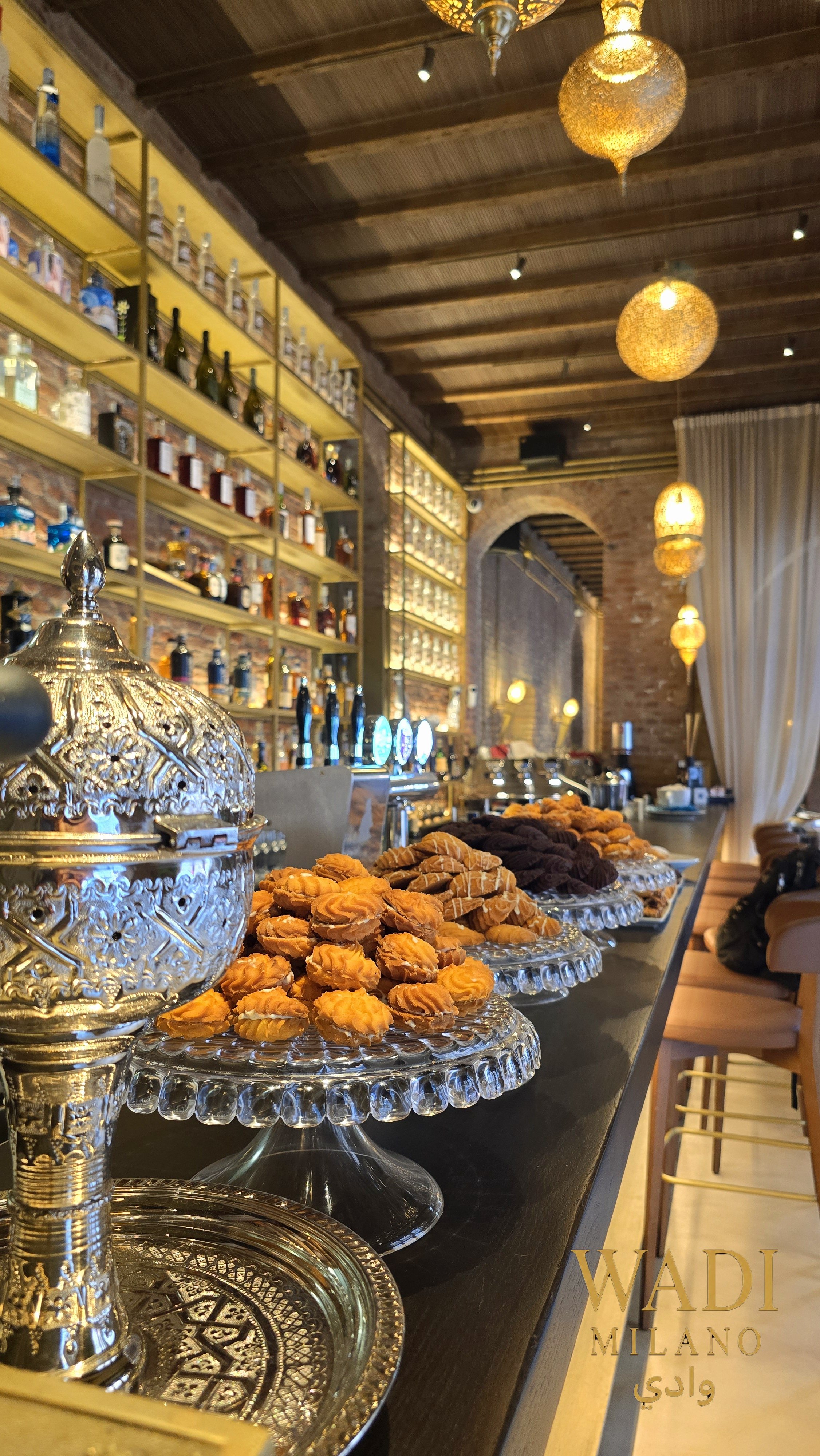 Interno di un ristorante con scaffali pieni di bottiglie di alcolici, illuminazione calda, lampade decorative sospese, tavolo con biscotti e dolci, tendaggi chiari, atmosfera accogliente e raffinata.