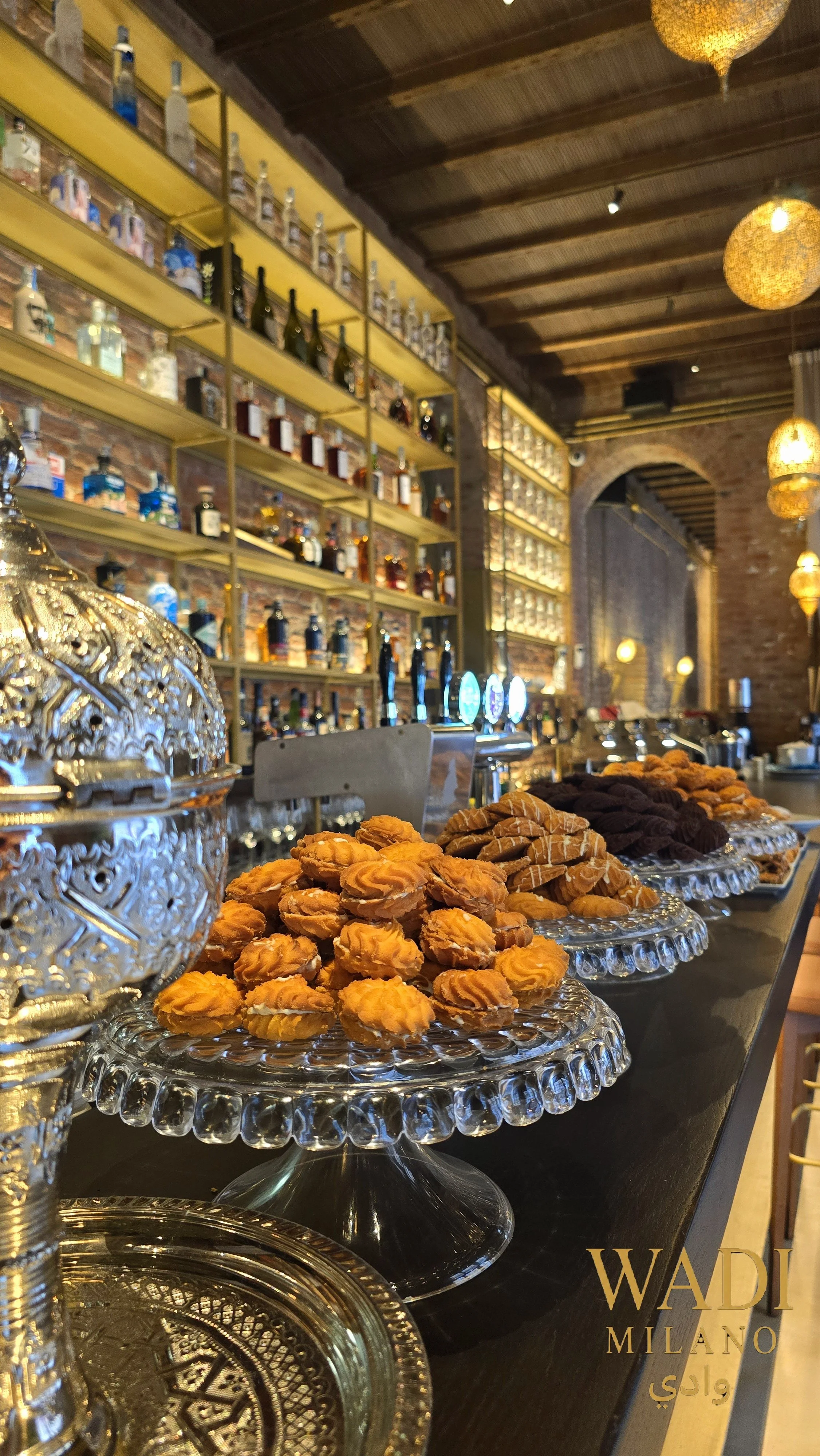 Vetrina di pasticcini e biscotti in un bar elegante con scaffali di bottiglie di alcolici sullo sfondo e luci calde