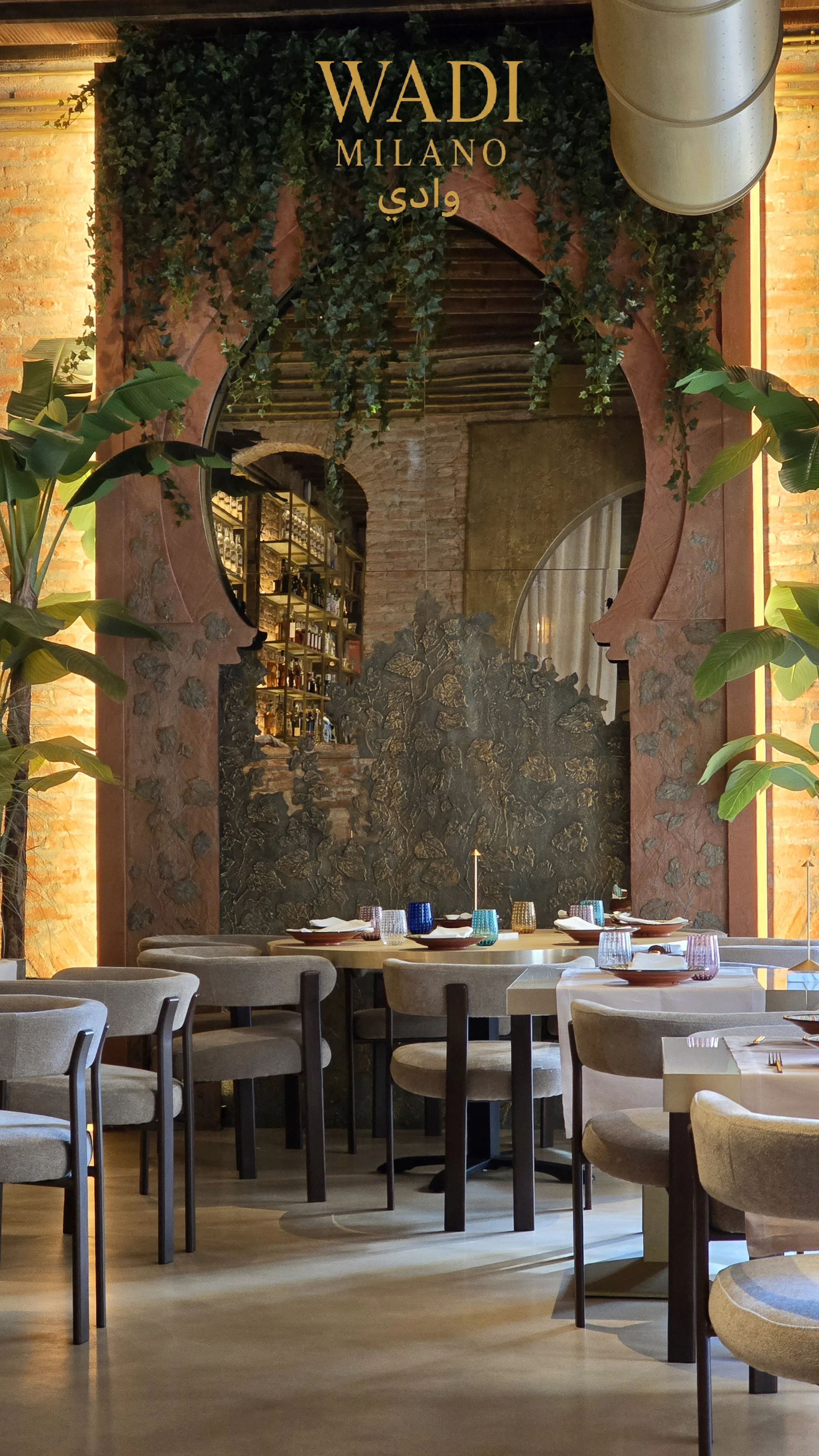 Interno di un ristorante elegante con tavoli arredati con bicchieri di vetro colorati e piatti, pareti di mattoni e decorazioni con piante verdi e specchi.