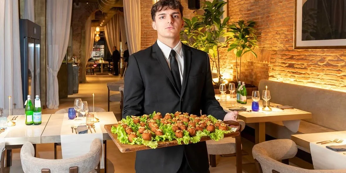 Uomo in abito nero che tiene un vassoio con antipasti in un ristorante elegante con ambiente caldo e tavoli apparecchiati.